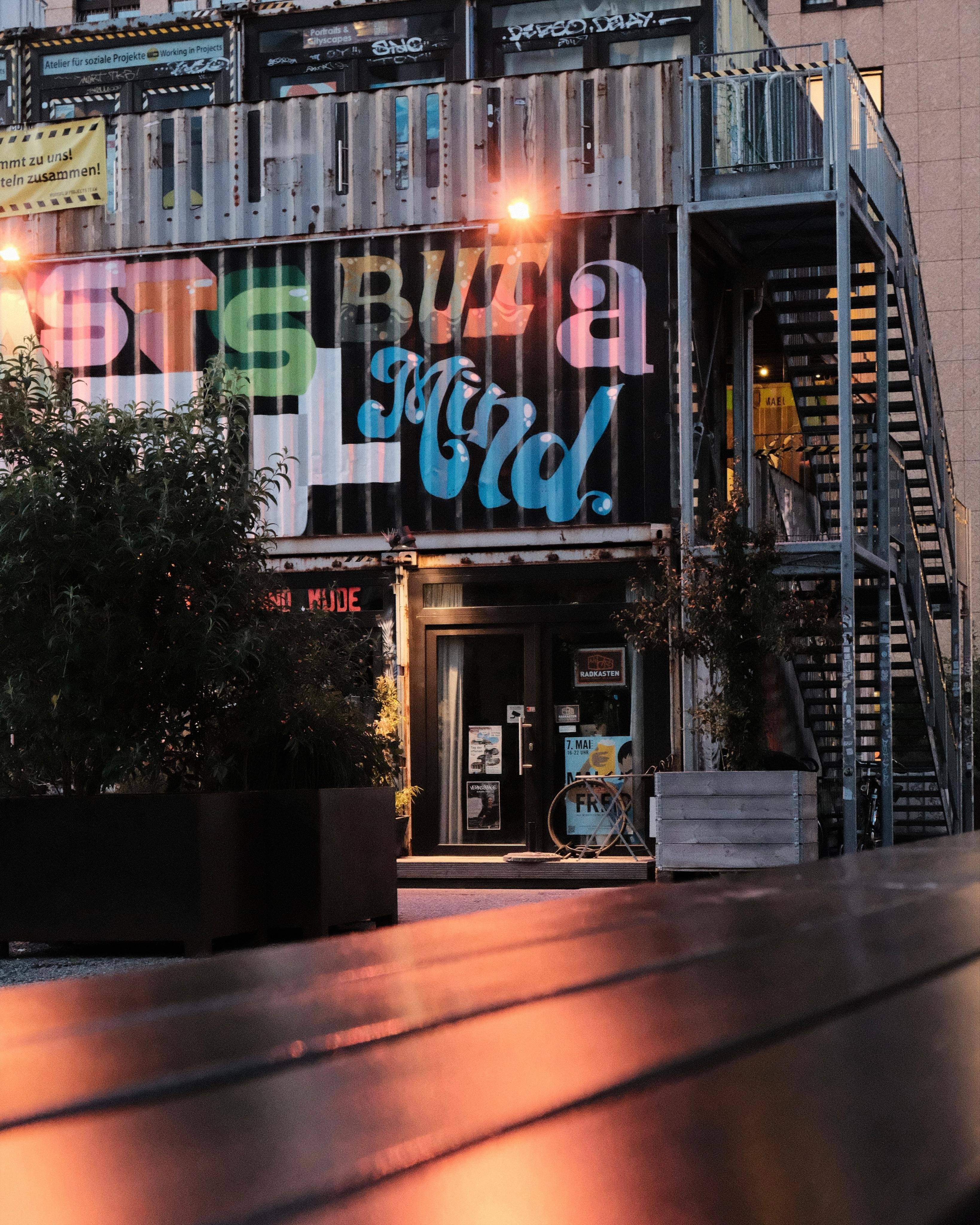 Two-story shipping-container building with large painted lettering on the upper wall, a ground-level entrance door with posters, and a metal exterior staircase on the right; planters and trees in front and a low bench in the foreground reflecting orange light at dusk.