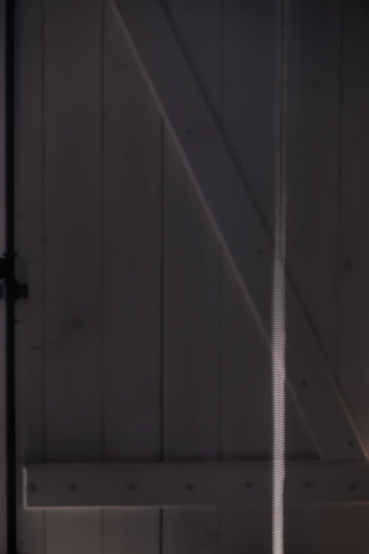 Photo verticale. Un volet en bois blanc presque totalement fermé et un raie de lumière frappant une surface invisible (une moustiquaire)