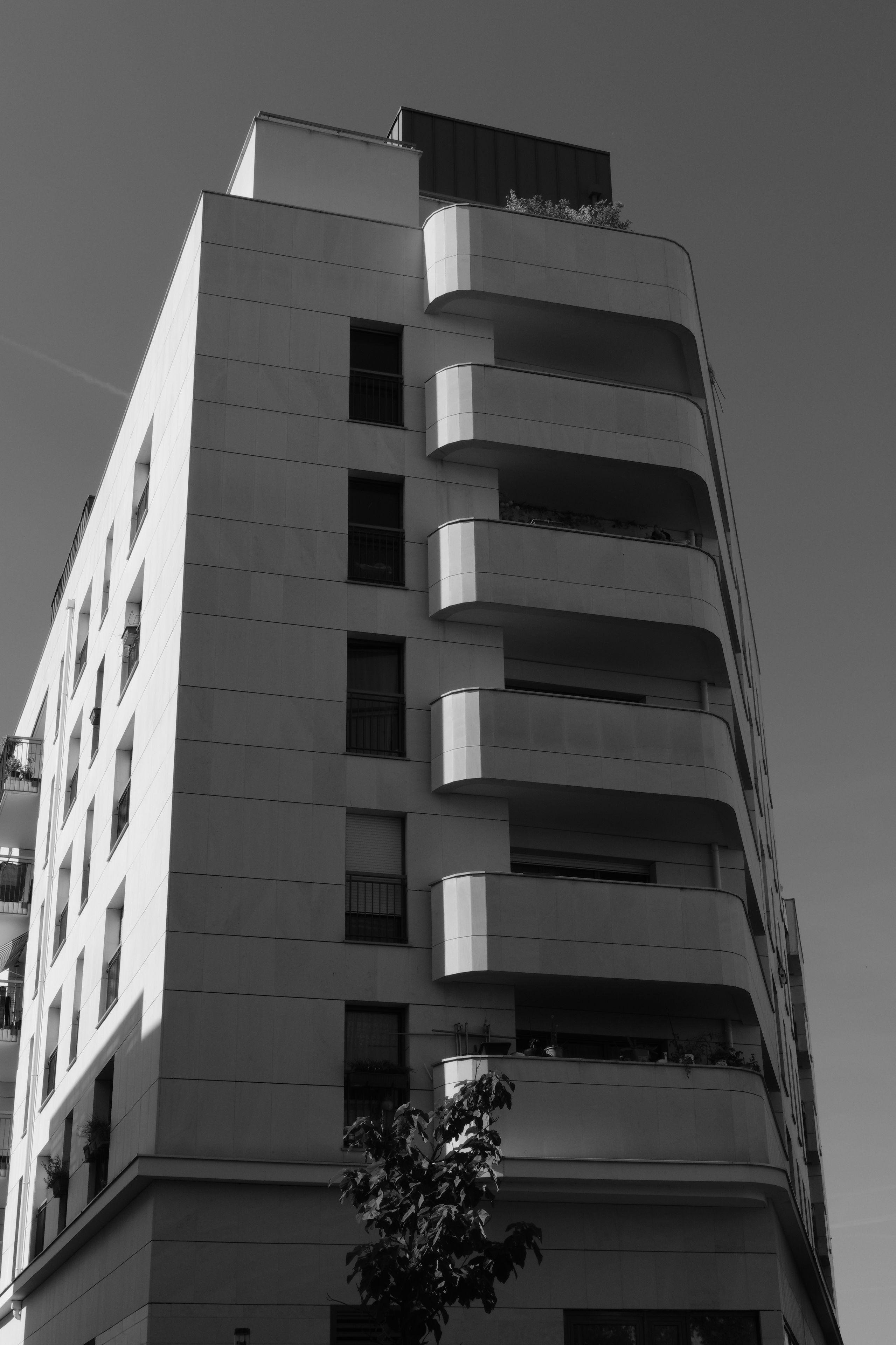 Black and white photo of a modern multi-story building with rounded balconies and some visible plants on the terraces, taken from a low angle. The sun lights the left side of the building