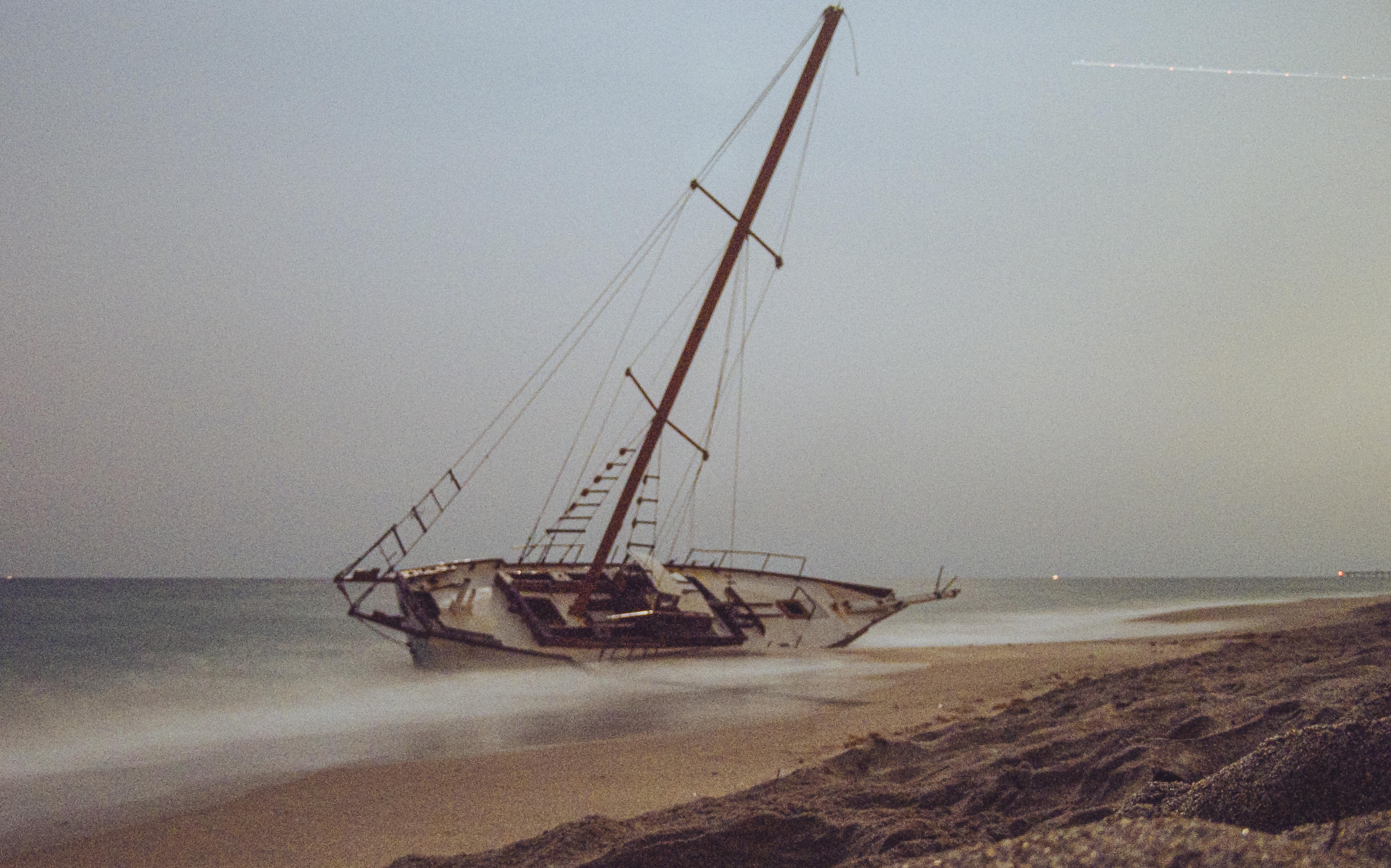 The image shows an abandoned sailboat leaning to one side on a sandy beach. The boat appears weathered and old, and it is positioned close to the shoreline with the sea in the background.