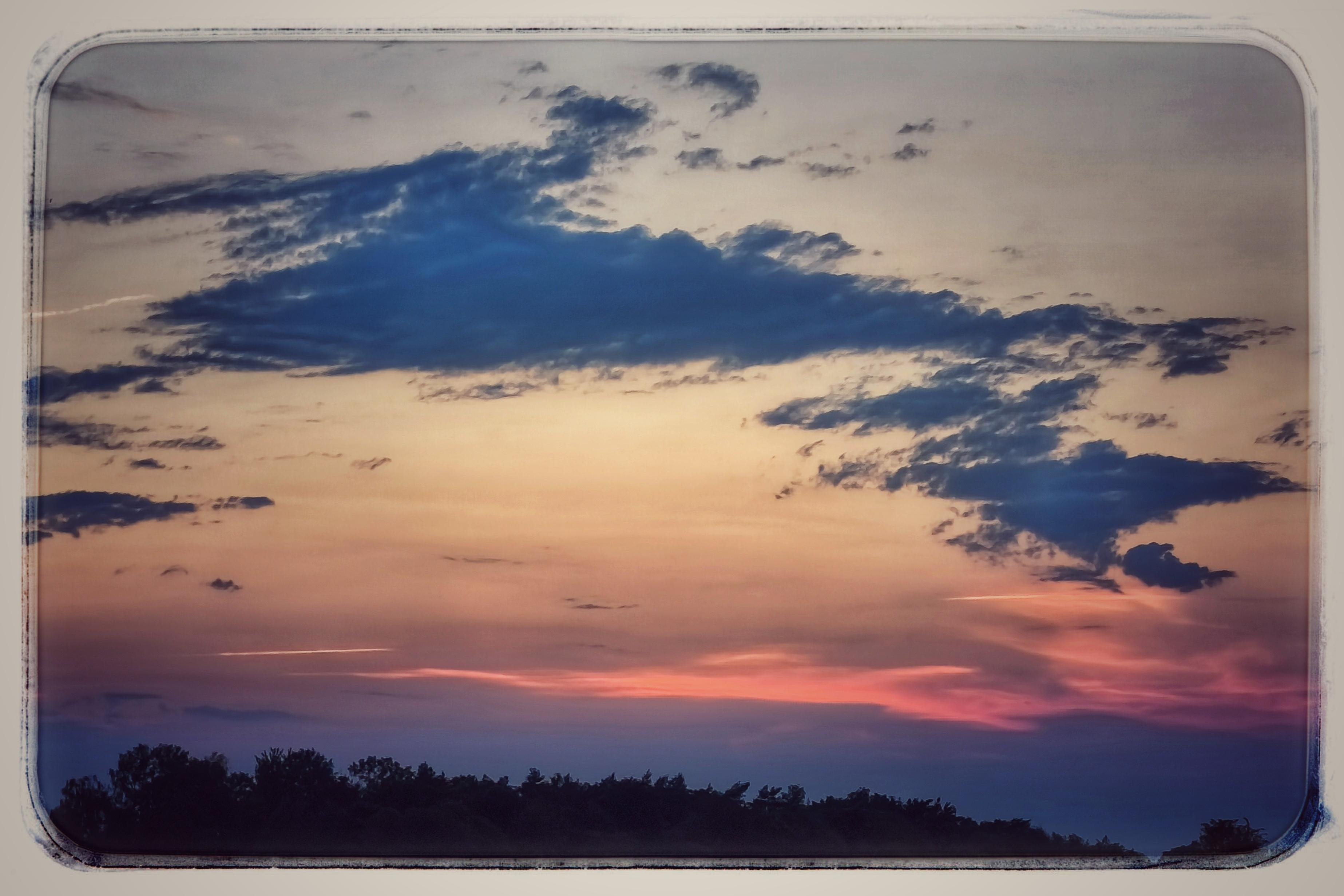 Wide sunset sky with dark blue clouds over a silhouetted tree line, fading from pale yellow near the horizon to pink and purple, framed by a rounded rectangular border.
###
Weiter Sonnenuntergangshimmel mit dunkelblauen Wolken über einer Baumsilhouette; die Farben verlaufen von einem blassen Gelb nahe dem Horizont hin zu Rosa und Violett, eingerahmt von einer abgerundeten rechteckigen Umrandung.