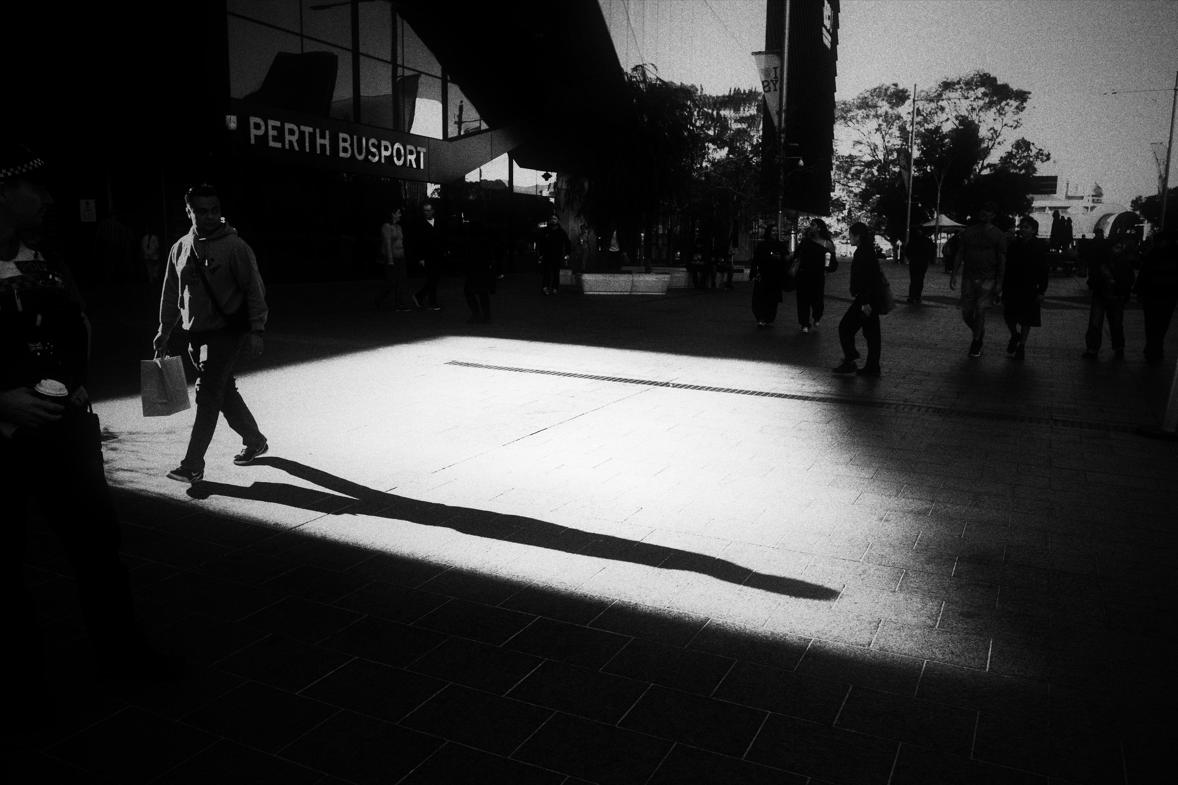 Black-and-white street scene outside a building marked “PERTH BUSPORT,” with a lone pedestrian carrying a bag crossing a bright sunlit patch of pavement, casting a long shadow, and several other people silhouetted in the darker background.