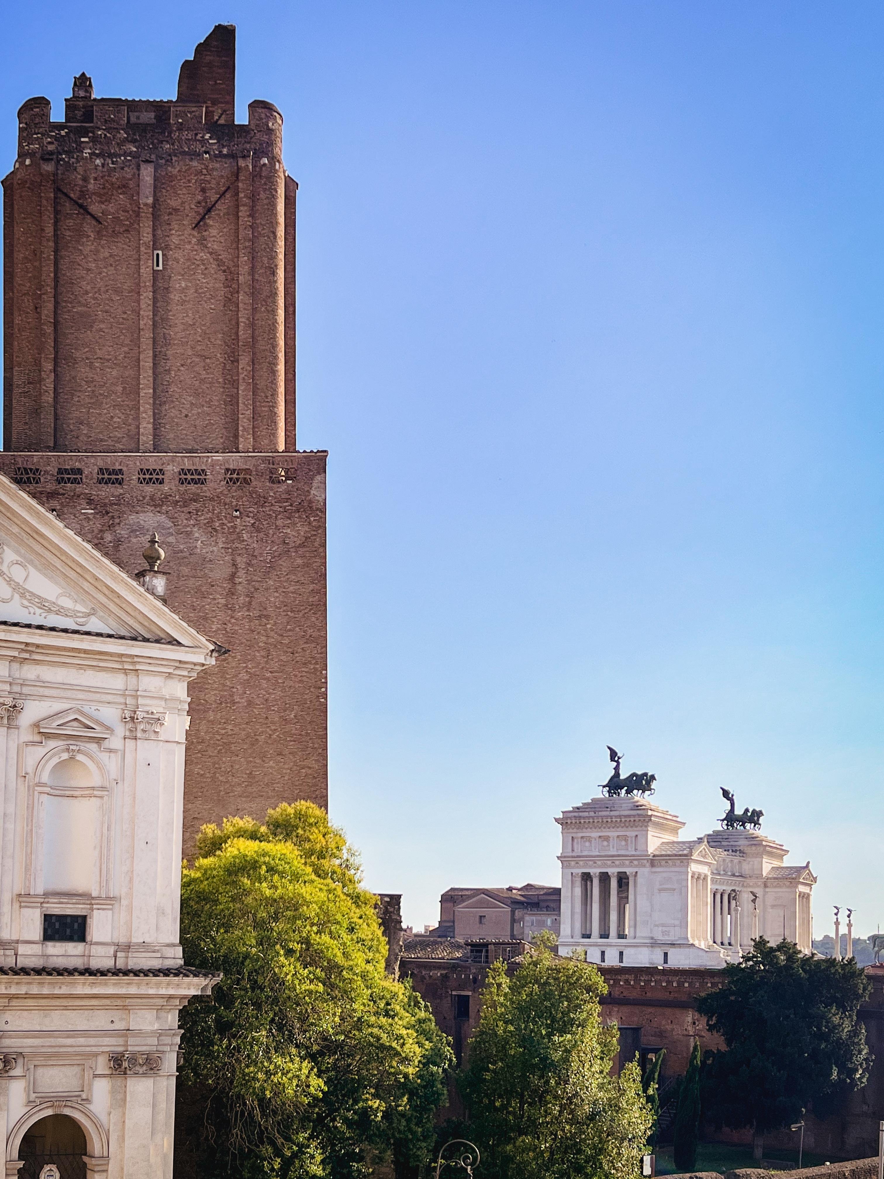 Paesaggio urbano con architettura storica. A sinistra c’è una torre alta e antica in mattoni chiamata "Torre delle Milizie". A destra, c’è parte di un edificio neoclassico bianco con statue sul tetto, chiamato Vittoriano. Il cielo è limpido e azzurro, e ci sono alberi in primo piano.