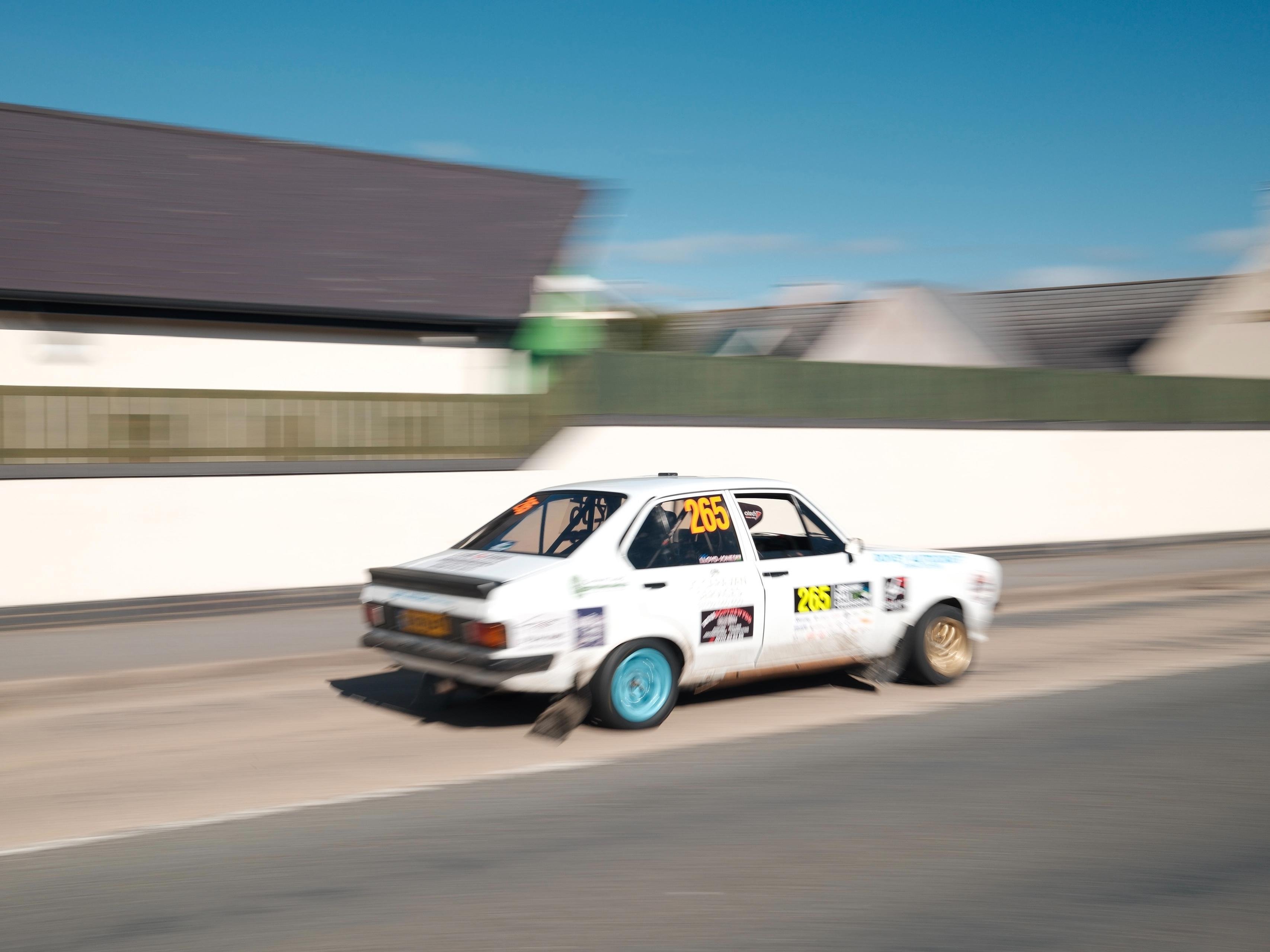 White rally car with number 265 on the side speeds along a paved road, motion-blurred, with a low wall and houses in the background under a blue sky.