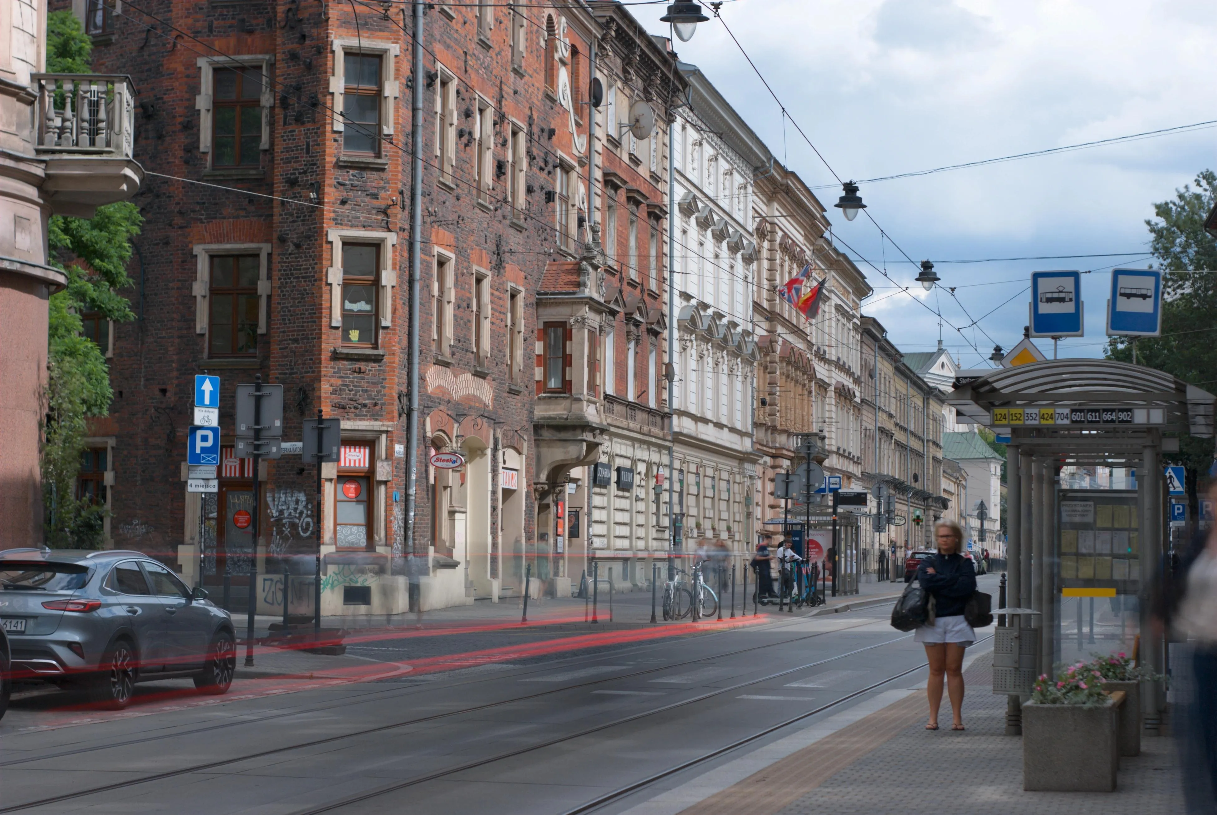 A photo of a person waiting for a bus and blurred car lights on the street during a day