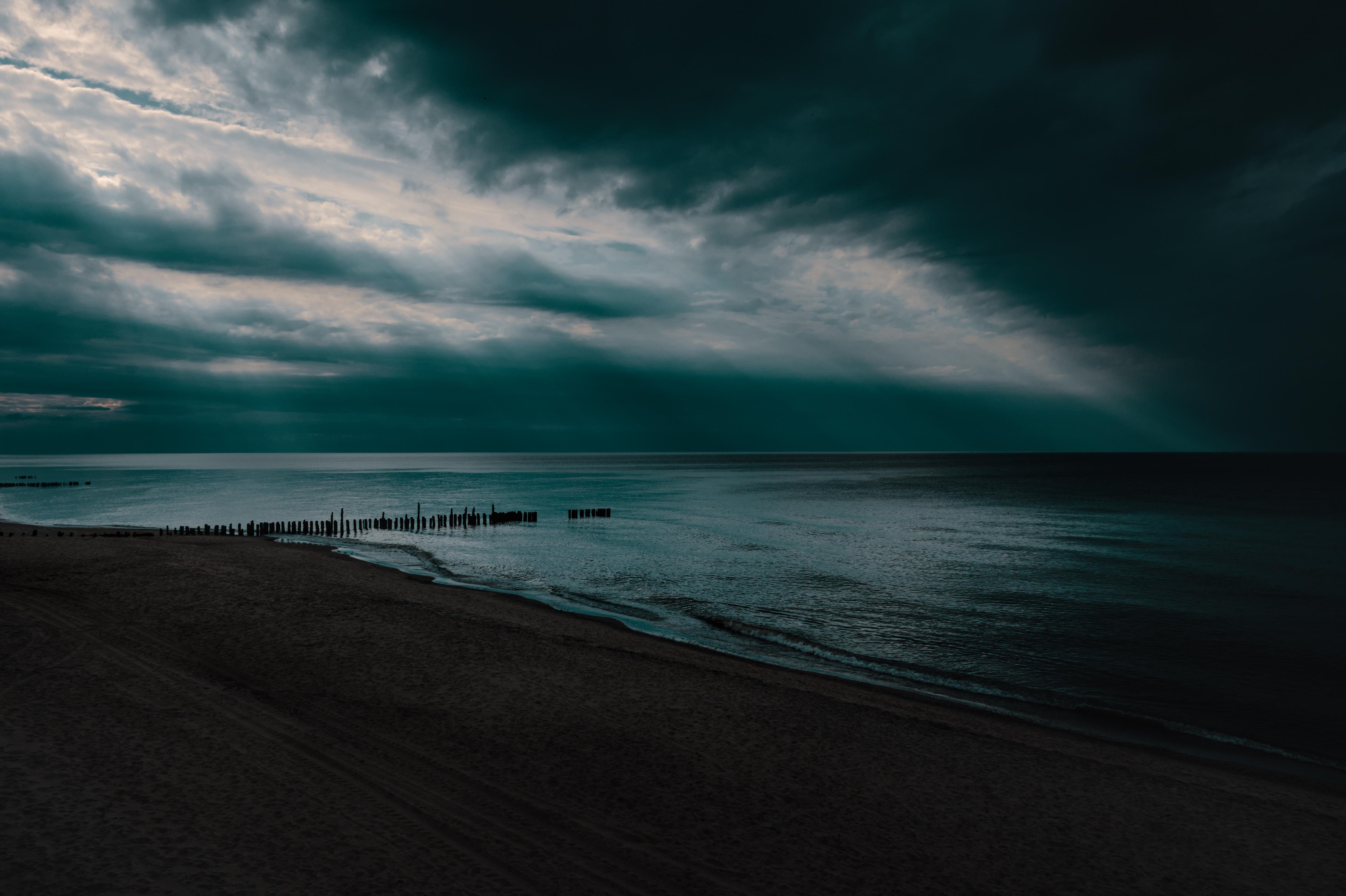 Dark sandy beach with a gentle shoreline, calm sea, and a line of wooden breakwater posts extending into the water under heavy, layered storm clouds with a bright opening near the horizon.