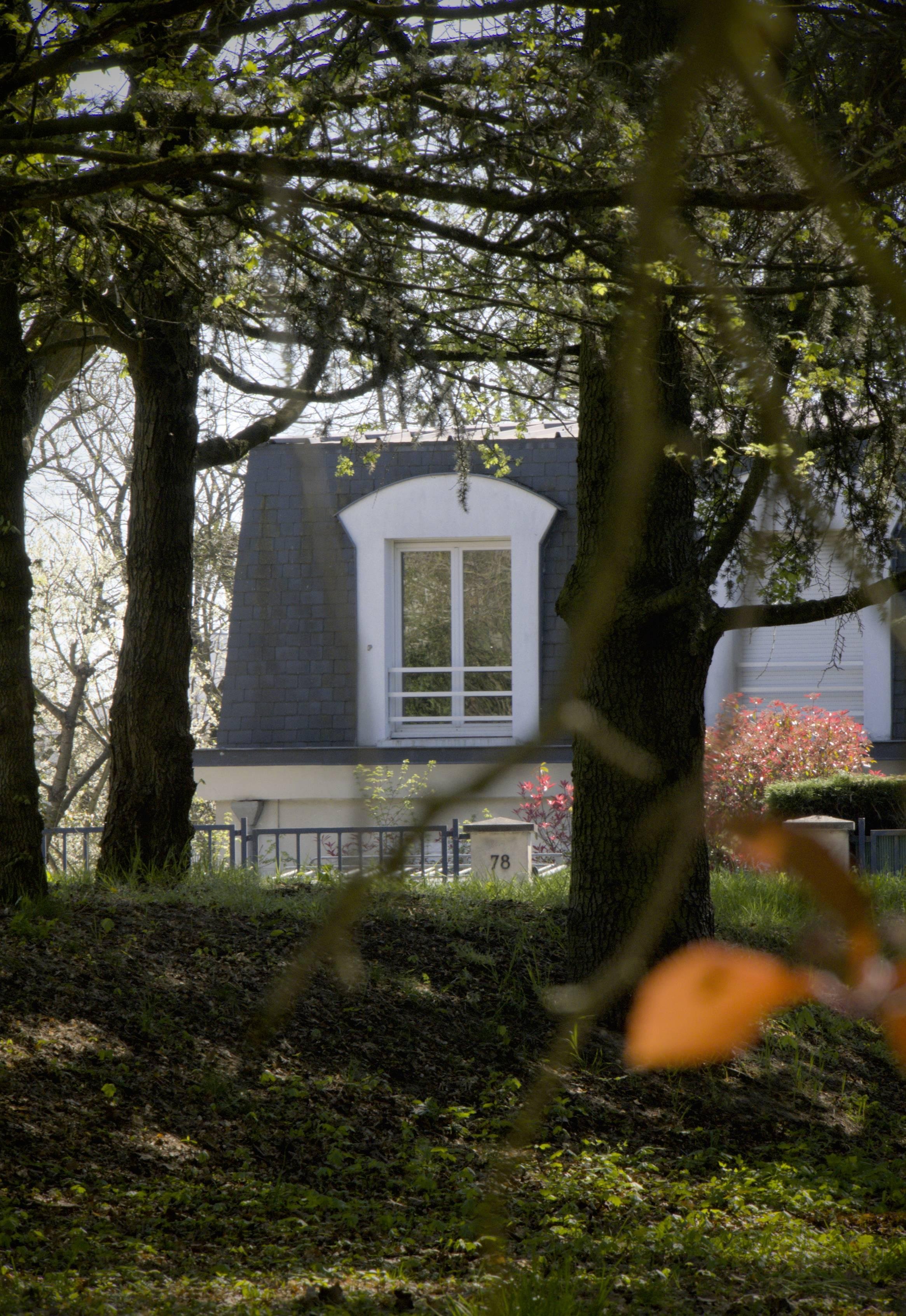 L'image montre une maison partiellement masquée par des arbres. La mise au point est faite sur une fenêtre surmontée d'un toit en ardoise. Au premier plan, des branches d'arbres et du feuillage sont flous, et à l'arrière-plan, une maison avec un numéro « 78 » sur un poteau près de l'entrée. Le cadre semble être une zone boisée avec de la lumière filtrant à travers les feuilles.