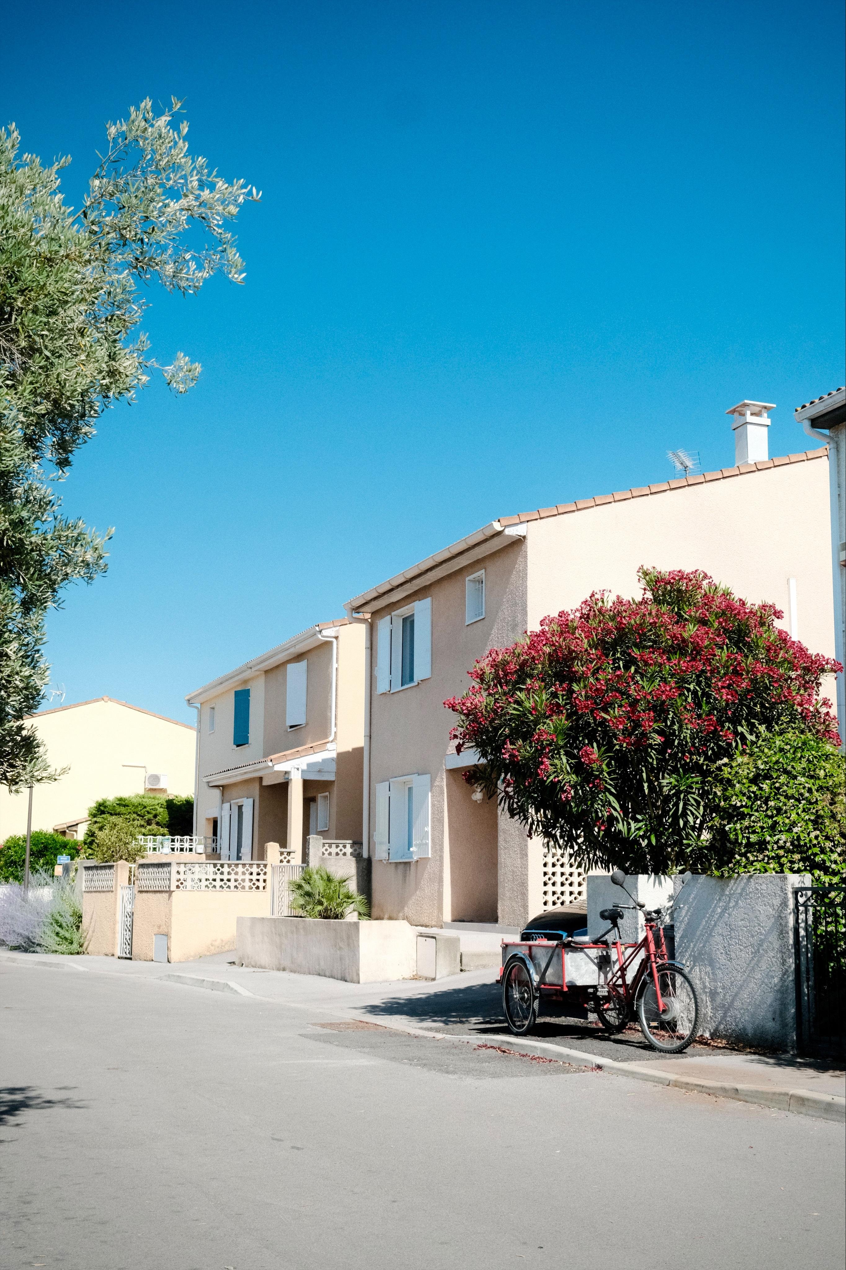 Residential street with beige two-story houses and white shutters under a clear blue sky, a flowering tree beside a low wall, and a red cargo tricycle parked at the curb in the foreground.