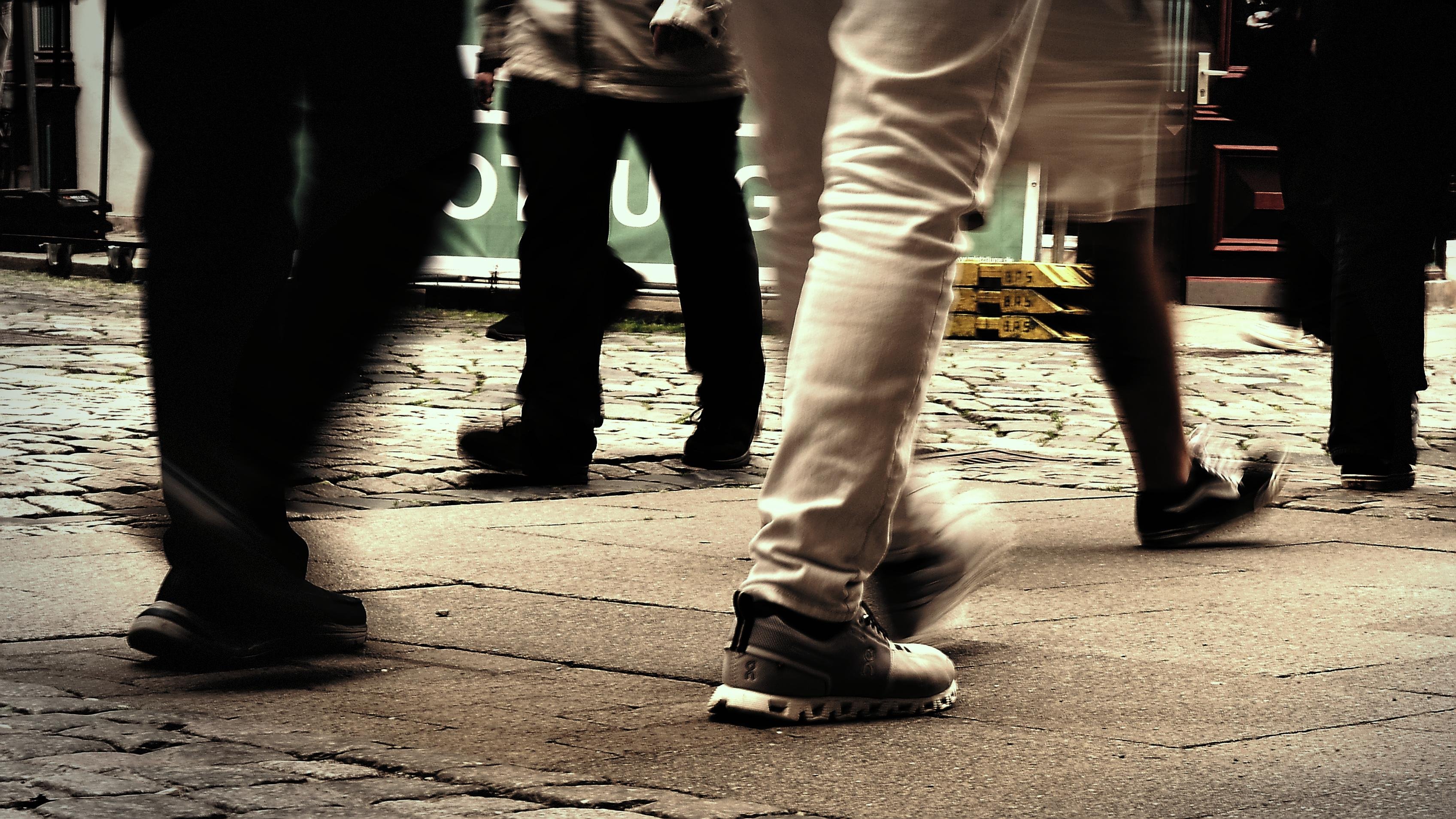 Low-angle street photo showing several pedestrians’ legs and shoes walking across a cobblestone and concrete sidewalk, with motion blur and storefront signs in the background.
###
Low-Winkel-Straßenfoto zeigt mehrere Fußgängerbeine und Schuhe, die über einen Kopfsteinpflaster und Beton-Bürgersteig gehen, mit Bewegungsunschärfe und Schaufensterschildern im Hintergrund.