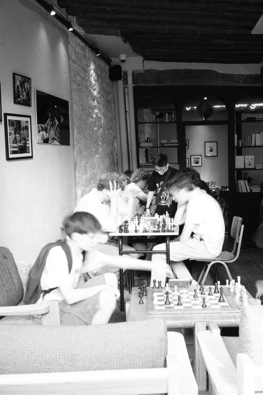 Photo verticale noir et blanc. Salle d’un bar à jeux où des partie d’échec sont en coures, le mouvement des mains floutés dans l’instant