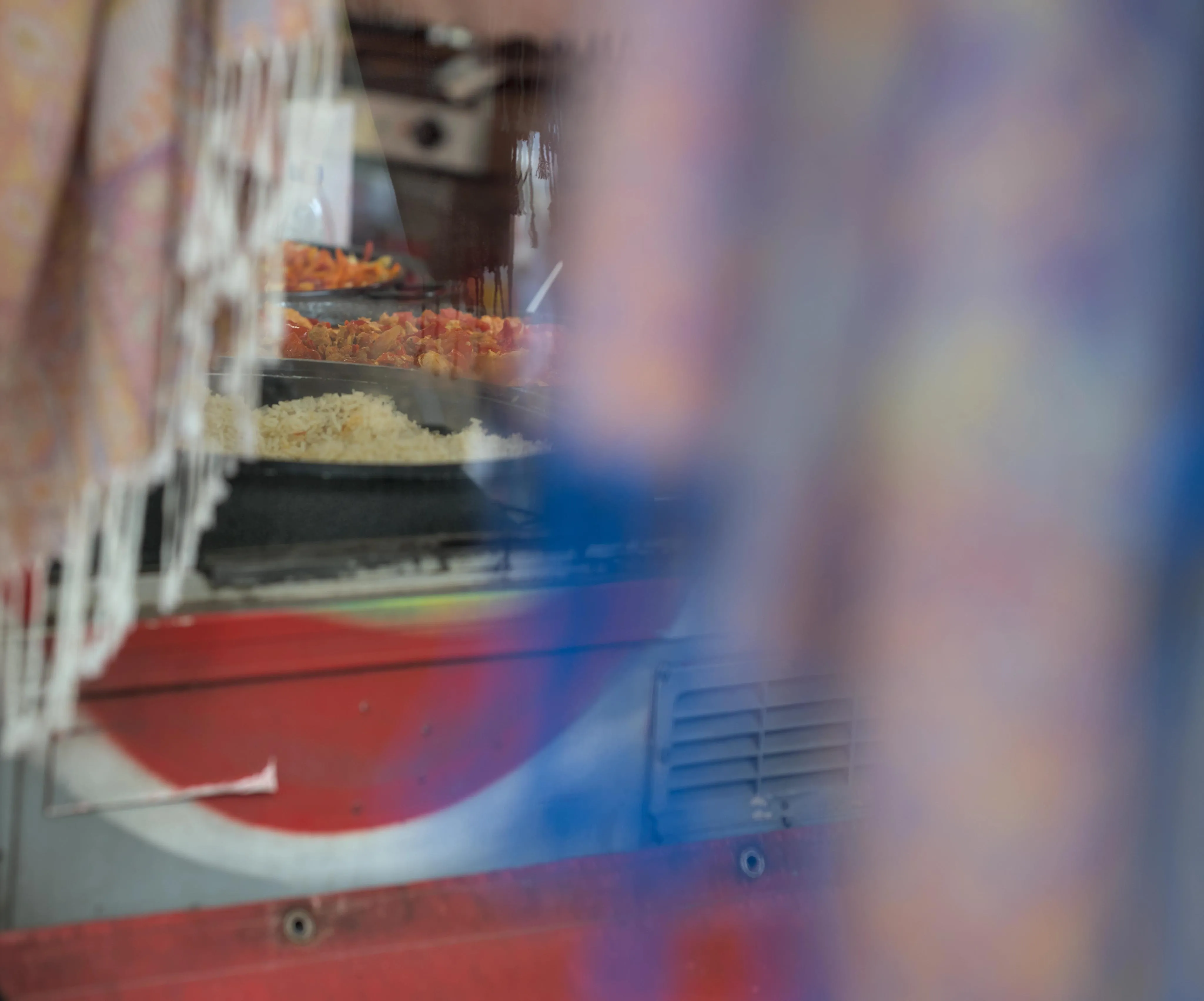 Close-up photograph on a market stall containing meals such as rice and meat with peppers, seen through a glass window. In the foreground blur, pieces of fabric with colored patterns hang on the left and right side of the image, forming a frame around the window into the food market stall.