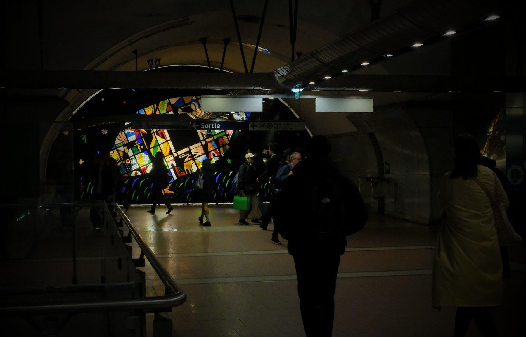 Photo horizontale. Un couloir de métro sombre, avec sur un mur au fond de la photo, un vitrail rétro éclairé. Des gens passent devant sans le voir