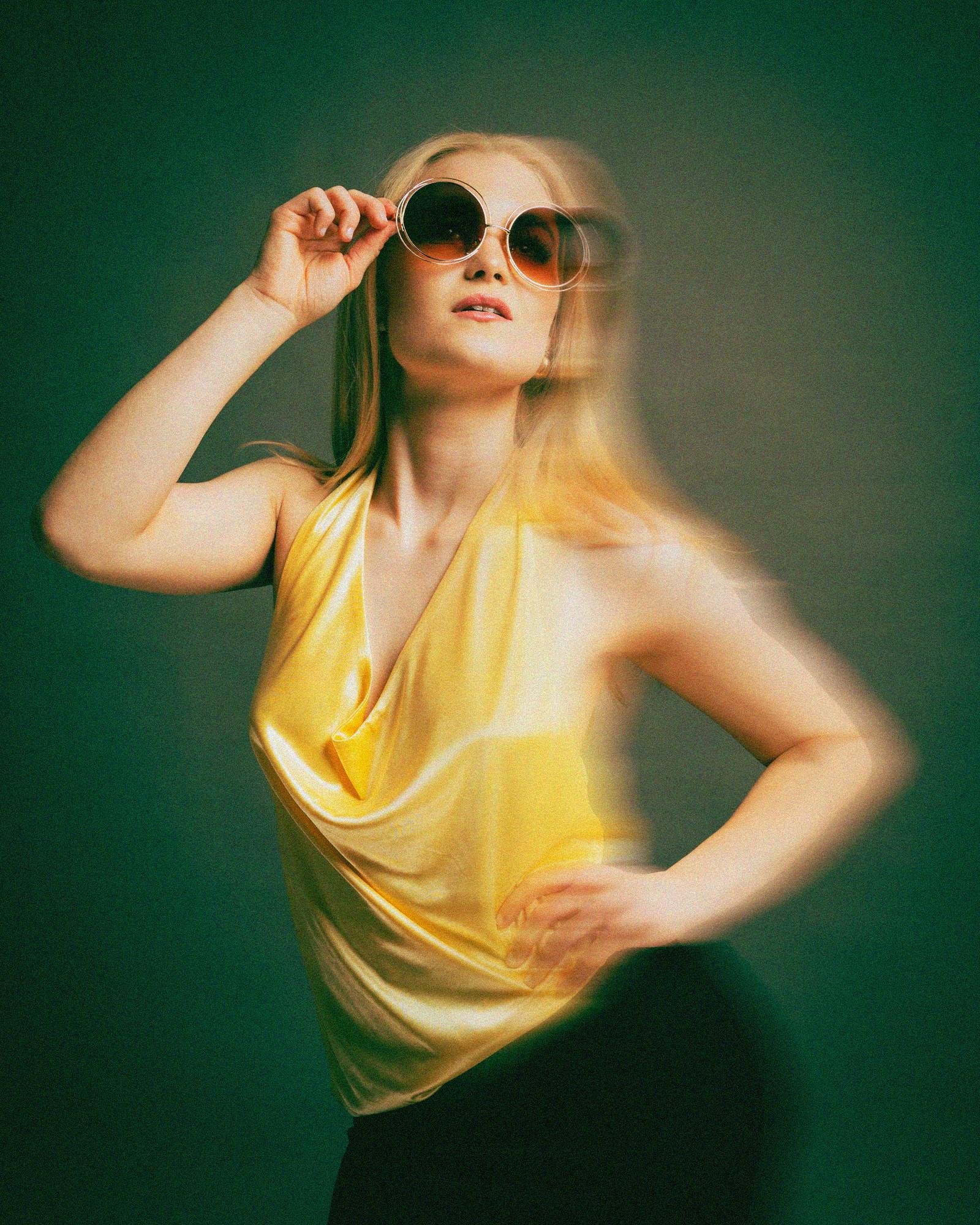 Person wearing large round sunglasses and a yellow halter top. They have long, light hair and are striking a pose with one hand on their hip and the other adjusting the glasses. The background is a soft, dark green. Motion blur adds a dynamic effect to the image.
