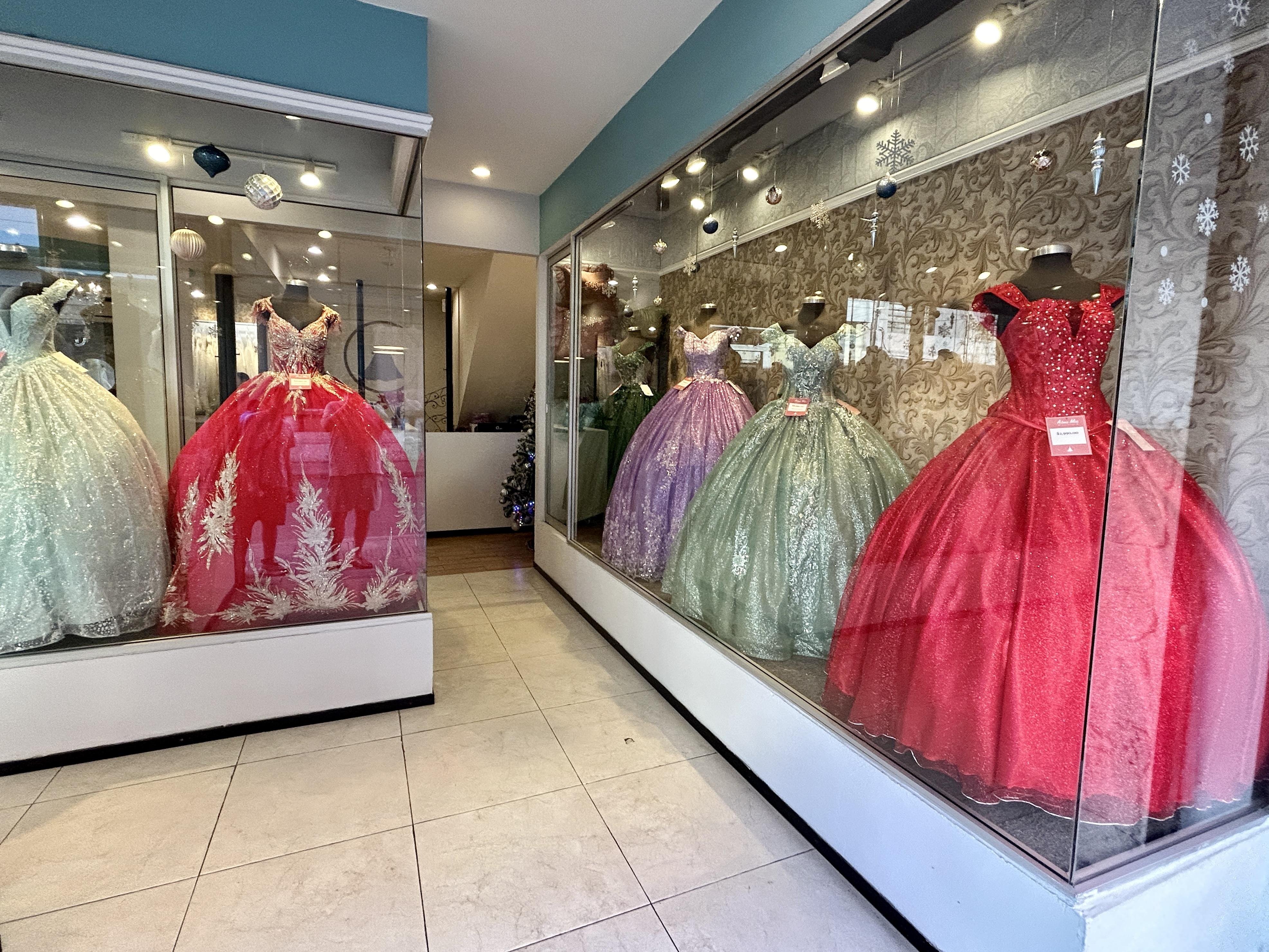 Glass display cases in a boutique holding six ornate ball gowns on mannequins—white and red dresses on the left, and purple, mint green, and two red dresses on the right—under spotlights with holiday snowflake decorations on the window; tiled floor and a hallway in the center.