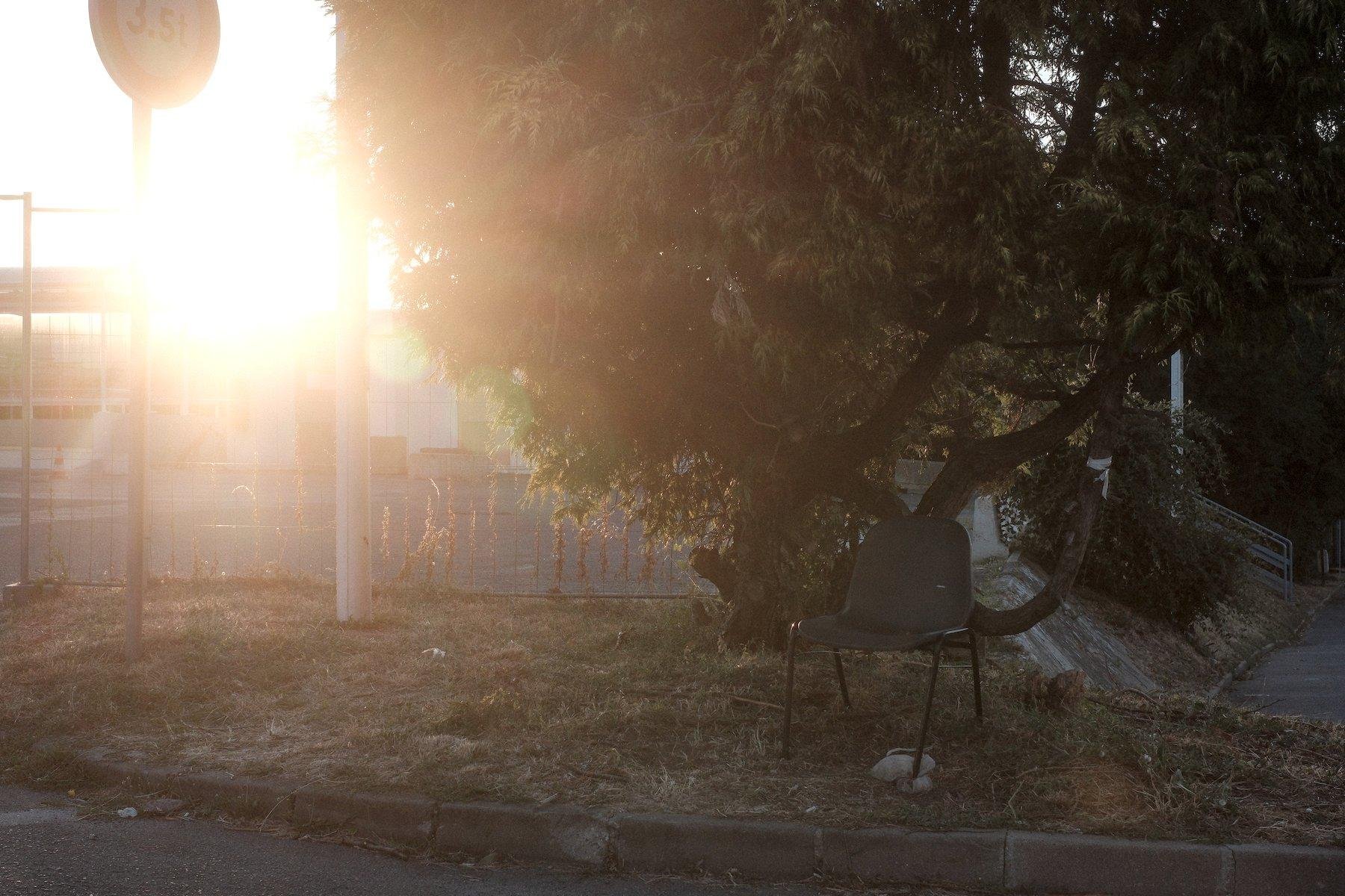 Photo horizontale. À droite de l’image, sous un petit arbre, une chaise grise a été posée sûrement pour avoir un endroit à l’ombre où se reposer. Le soleil couchant nous éblouis à gauche de l’image