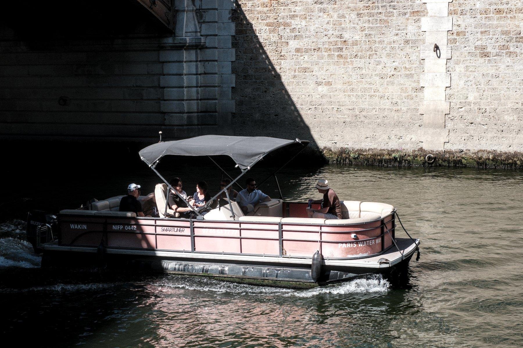 Photo horizontale. Un petit bateau de loisir sur la Seine avec des banquettes sur tout le pourtour, une grande ombrelle rectangulaire, sortant de l’ombre d’un pont. Des gens sont assis sur les banquettes et discutent