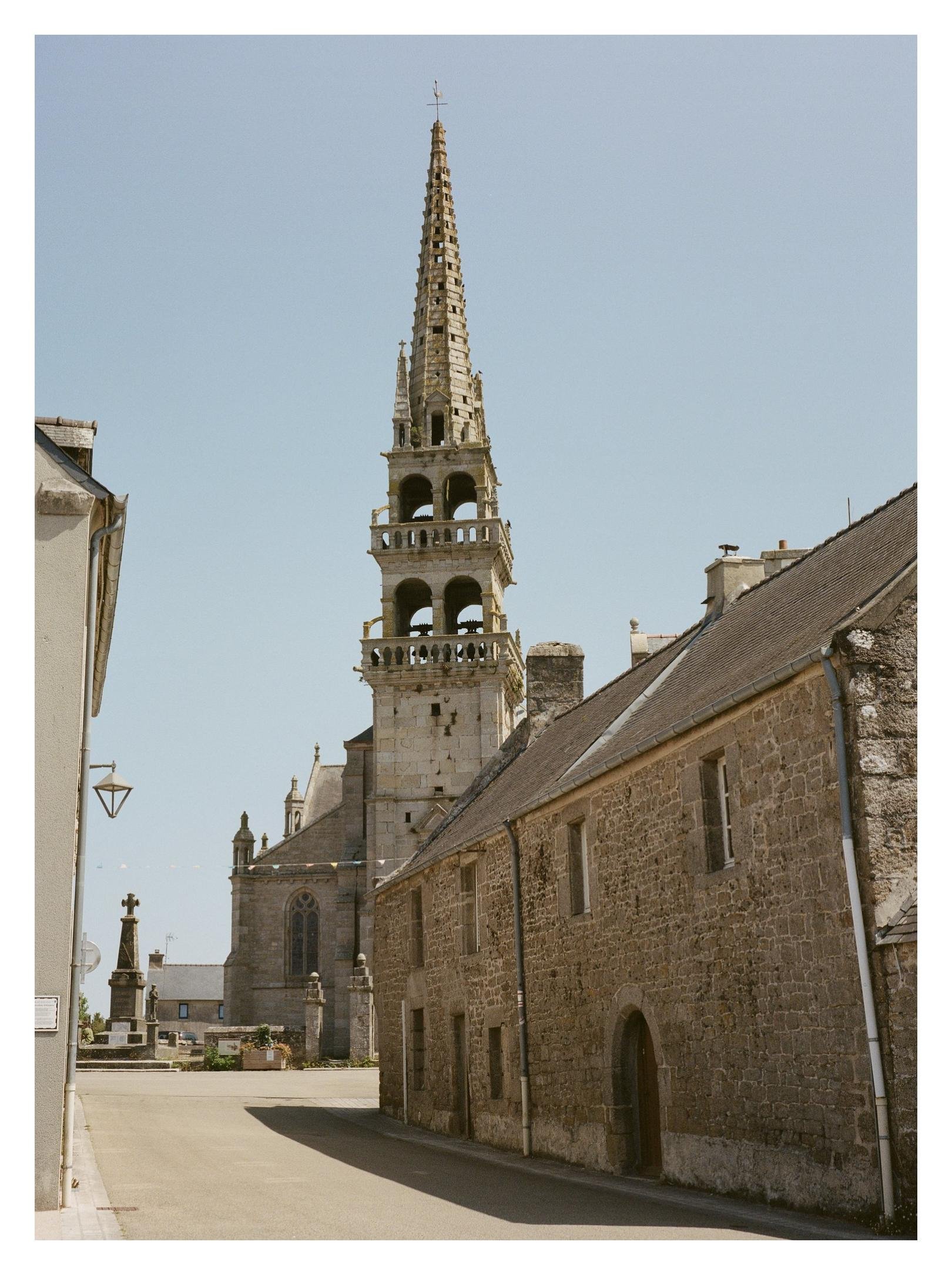 Tall stone church bell tower with a pointed spire rising above a village street, flanked by stone buildings and rooftops; a small monument stands in an open square under a clear blue sky.
