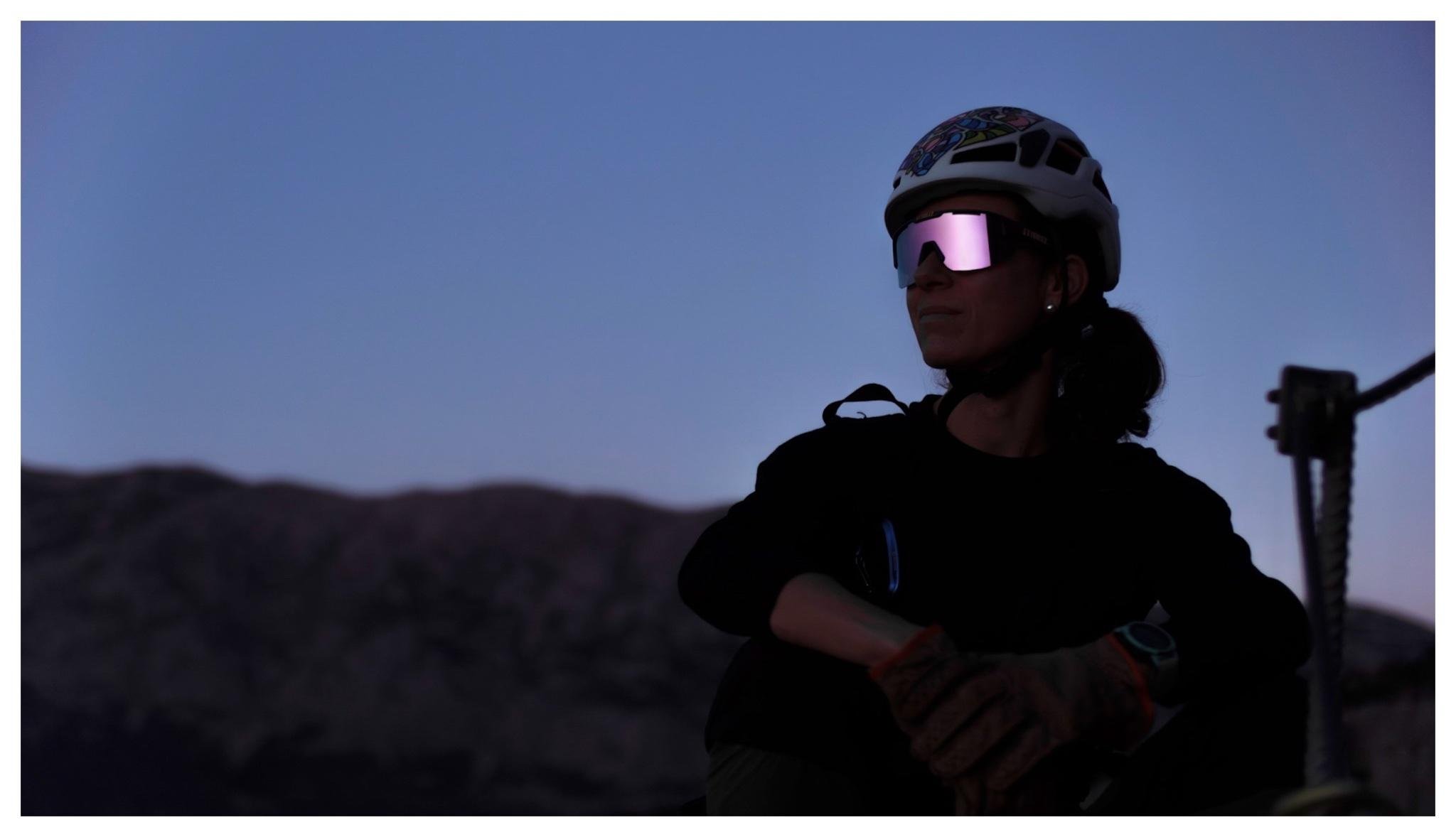 Person wearing a climbing helmet and reflective sunglasses sits with arms resting on knees in silhouette against a twilight sky, with dark mountain ridges in the background and a metal post on the right edge.