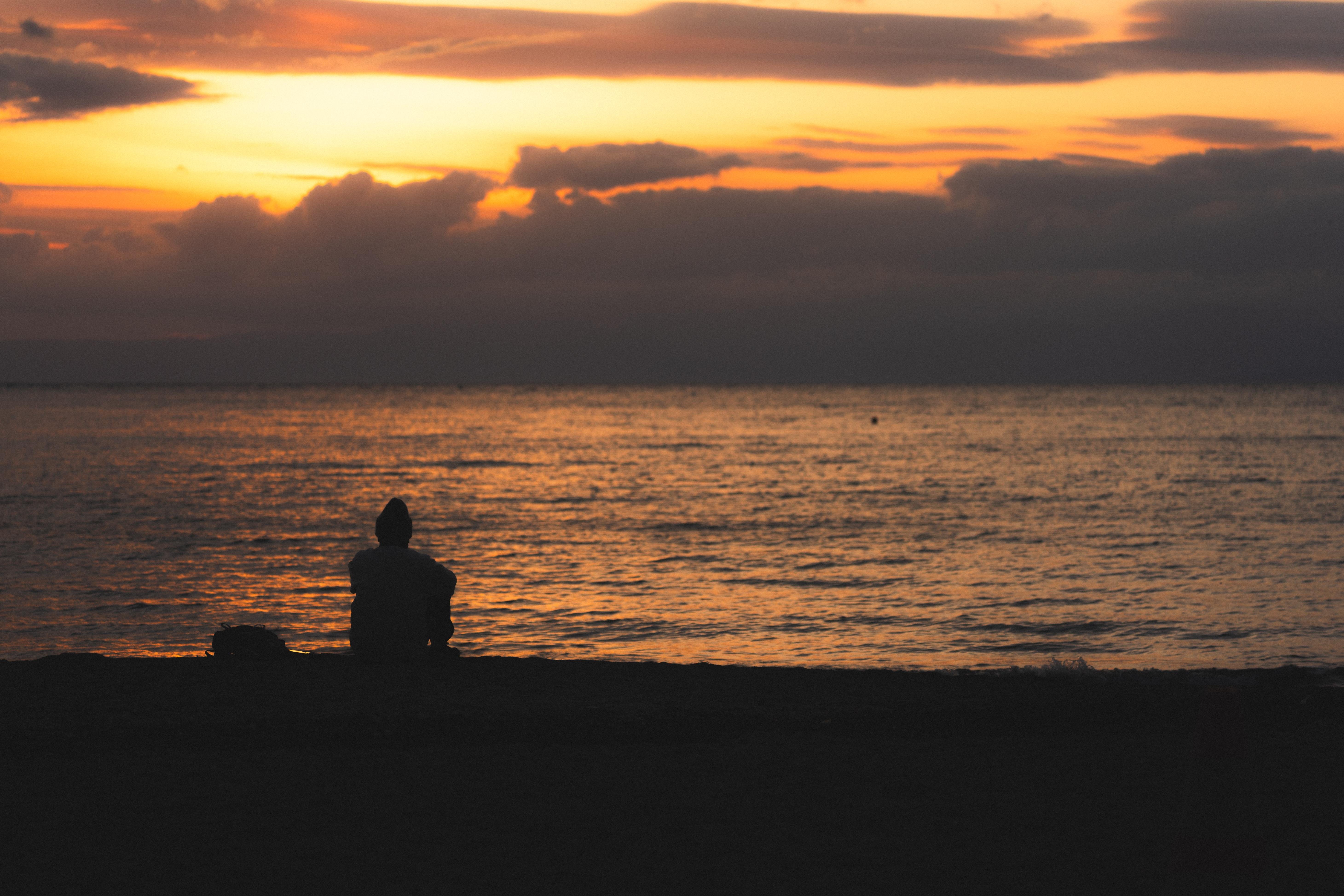 Silhouette of a person sitting on the beach facing the ocean at sunset. The sky features vibrant orange and yellow hues with scattered dark clouds. The ocean reflects the colors of the sunset, and the horizon is indistinct against the illuminated sky.