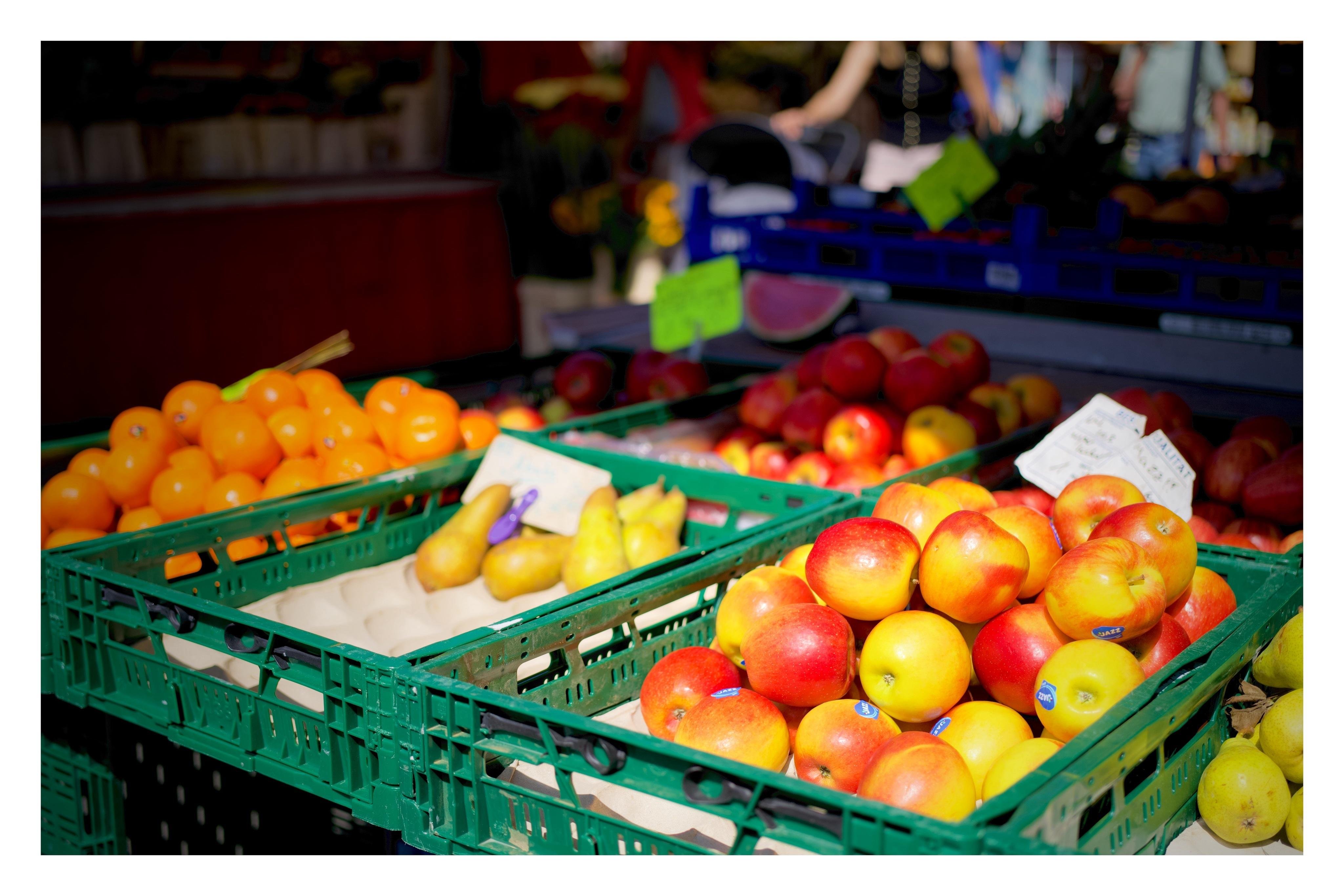 Frisches Obst auf einem Marktstand, darunter Äpfel, Birnen und Orangen in grünen Kisten; sonnige, farbenfrohe Szene im Freien.