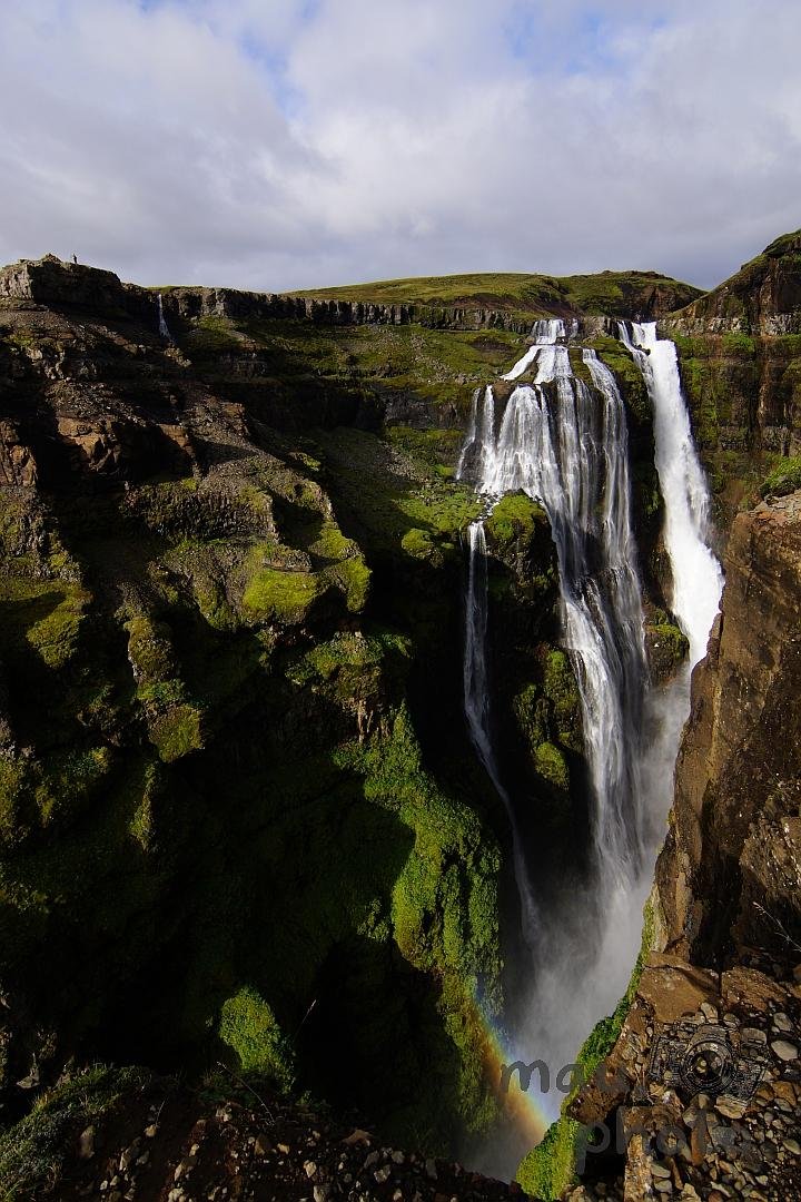 The image shows a stunning waterfall cascading down a rocky cliffside, surrounded by lush green vegetation. The water is flowing powerfully and there is a visible rainbow near the base where the mist accumulates. The sky is mostly cloudy, providing a dramatic backdrop to the scene.