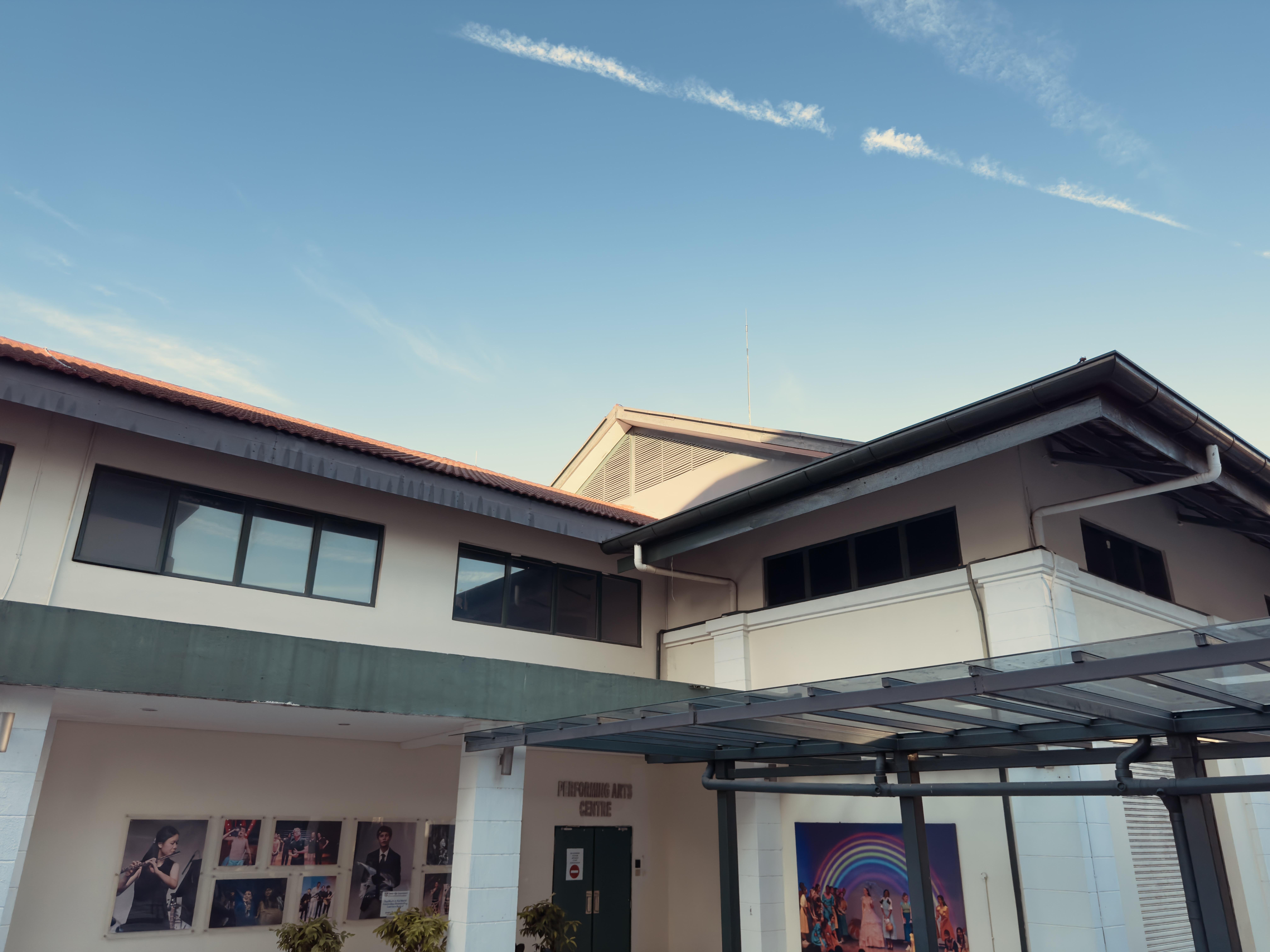 This is an image of a building labeled “Performing Arts Centre.” The structure appears to be modern, with large windows and a glass awning at the entrance. There are posters or images of performers on the wall, and the sky above is clear with a few clouds.
