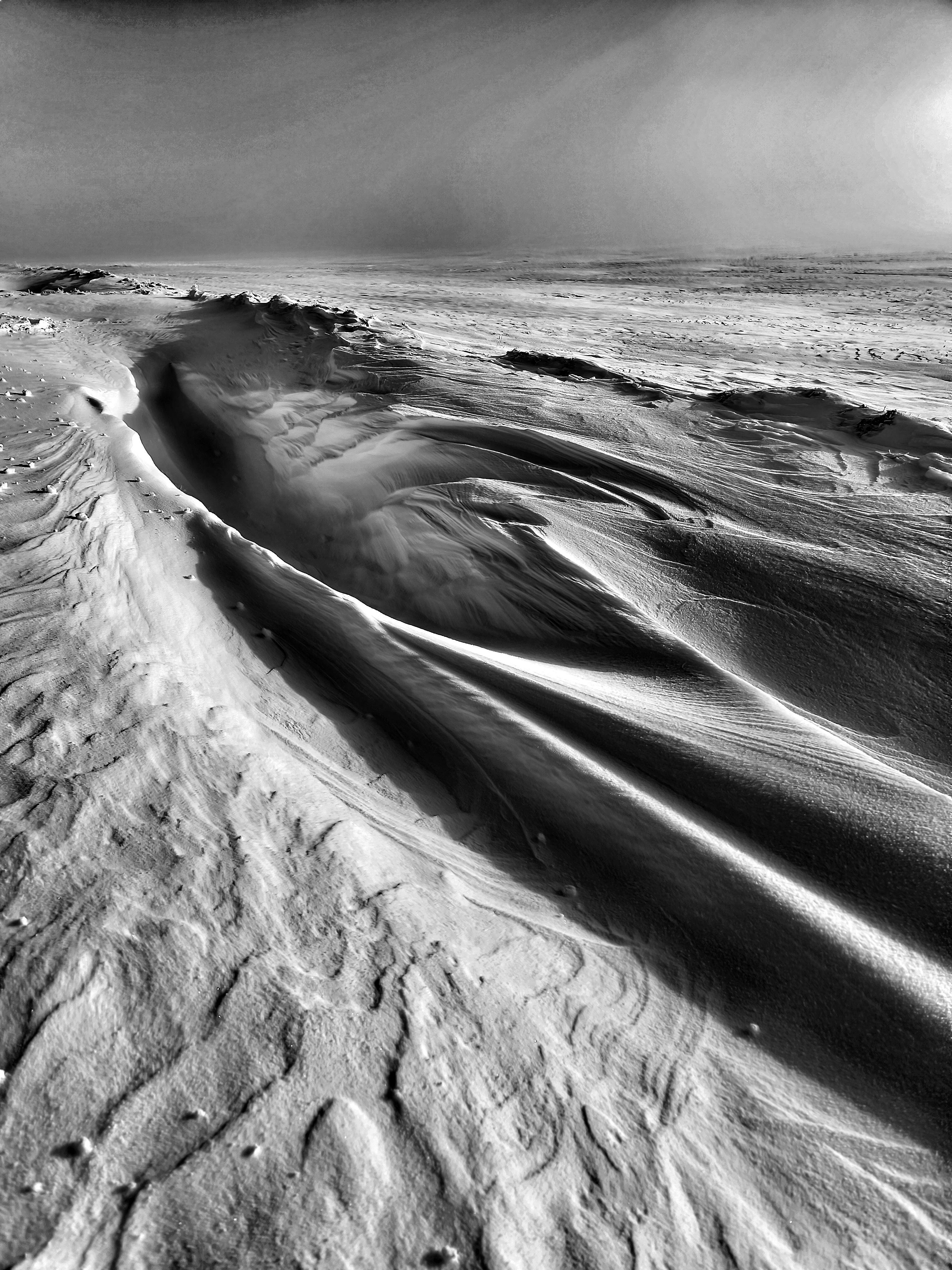 Black-and-white landscape of wind-sculpted snow dunes, with a sharp ridge running diagonally from the lower left toward the center and rippled textures in the foreground fading into a flat horizon under a bright, hazy sky.
###
Schwarz-weiße Landschaft aus vom Wind geformten Schneedünen, mit einem scharfen Grat, der diagonal von links unten zur Mitte verläuft, und gewellten Texturen im Vordergrund, die unter einem hellen, dunstigen Himmel in einen flachen Horizont übergehen.