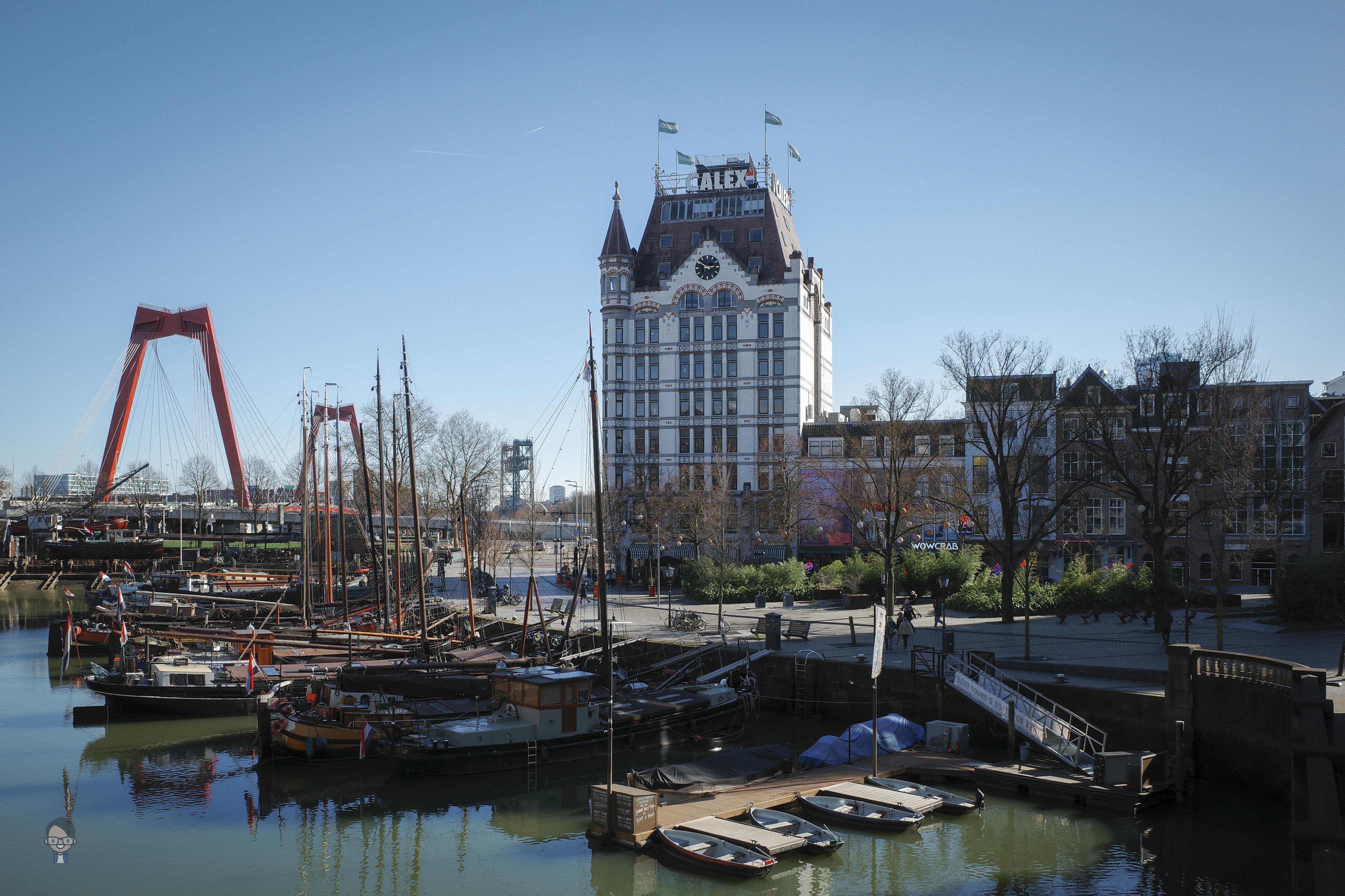 The image depicts a harbor scene with several docked boats. In the background, there is a tall, historic-looking building with distinctive architecture and a name on top. To the left, a red bridge can be seen, adding to the urban landscape. The area is surrounded by additional buildings and trees, with clear skies above. This could be a place of interest in a city, possibly used for tourism or historic exhibitions.