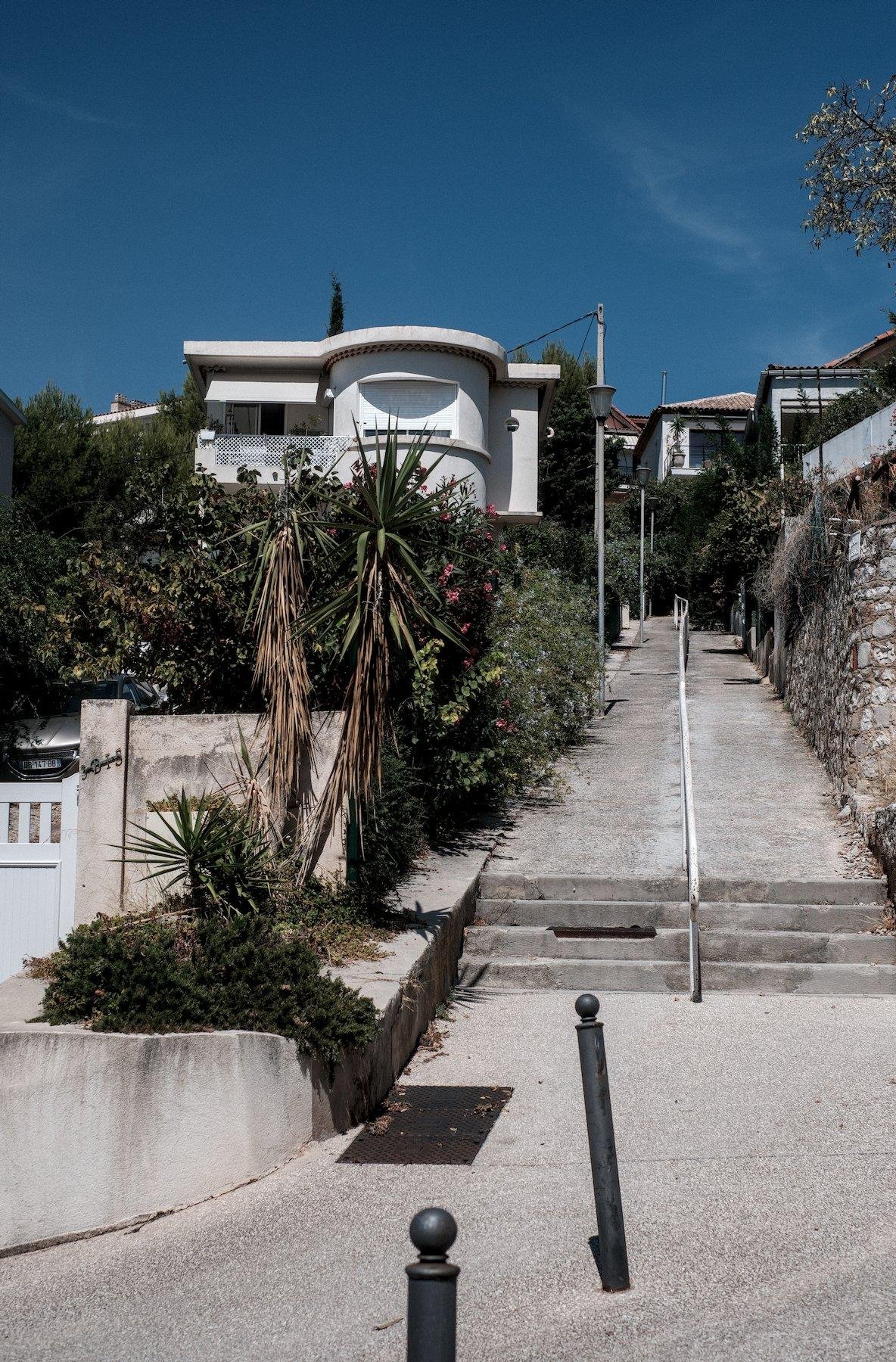Vertical photo. A cemented footpath curving into 4 steps and a quite long slop going up with a handrail at the center. A house in present on the left side with a modern architecture