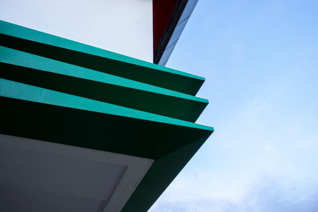 Green geometric architectural element with three horizontal slats extends from a white wall against a blue sky.