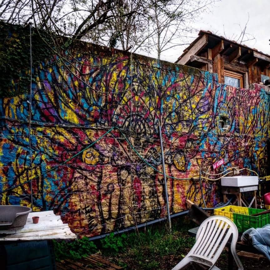 This image shows a colorful graffiti wall with abstract patterns and shapes, featuring a mix of red, yellow, blue, and black. In front of the wall, there's an outdoor setup with various objects, including a white plastic chair, a sink, a table with pots, and a green crate. The area has a casual and artistic vibe, and some trees are visible above the wall.