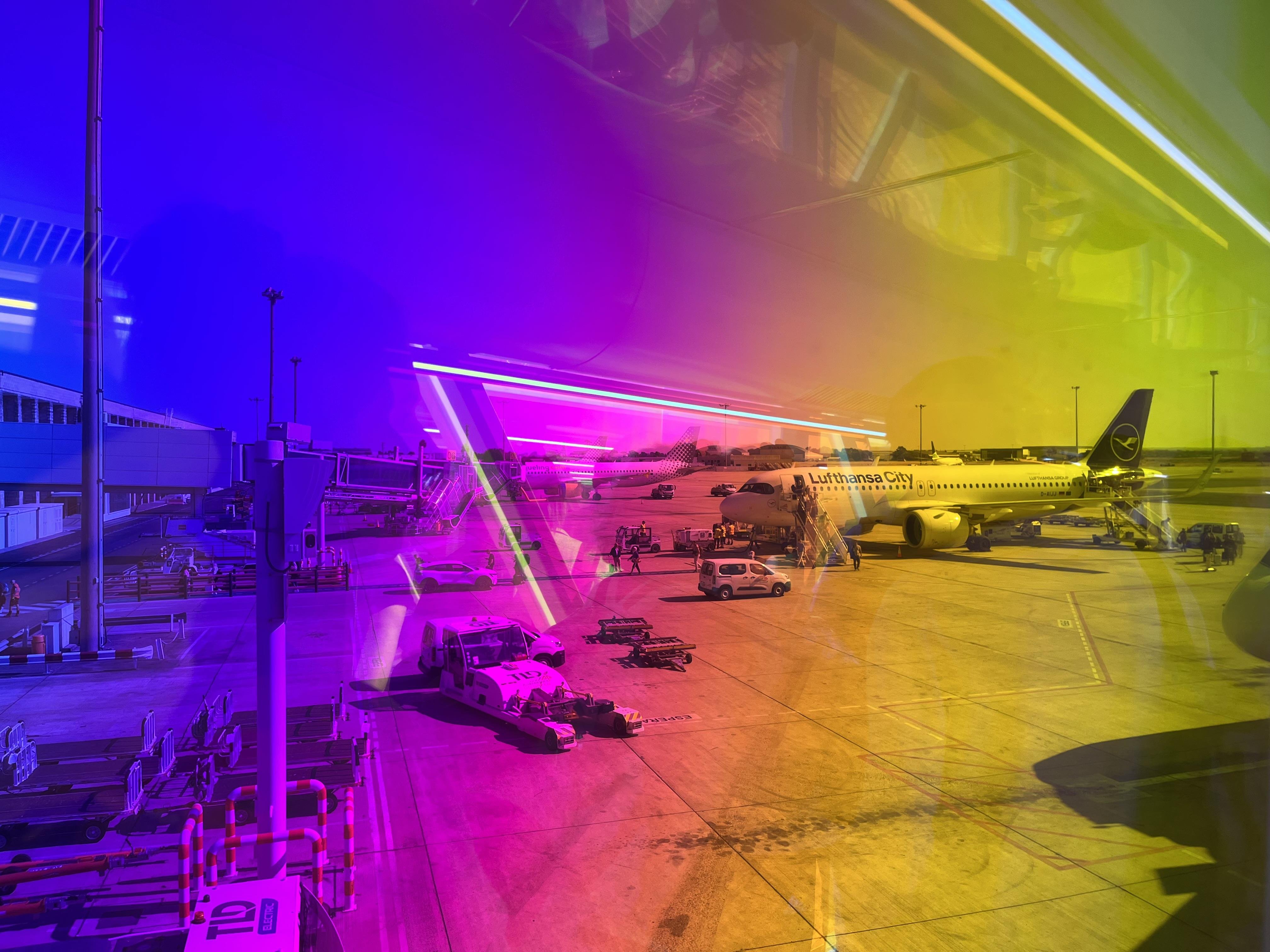 Airport gate apron viewed through tinted glass, with an aircraft parked at the terminal and several ground service vehicles and baggage carts nearby; purple-to-yellow color cast overlays the scene due to the foil covering the glass of the airport window.
