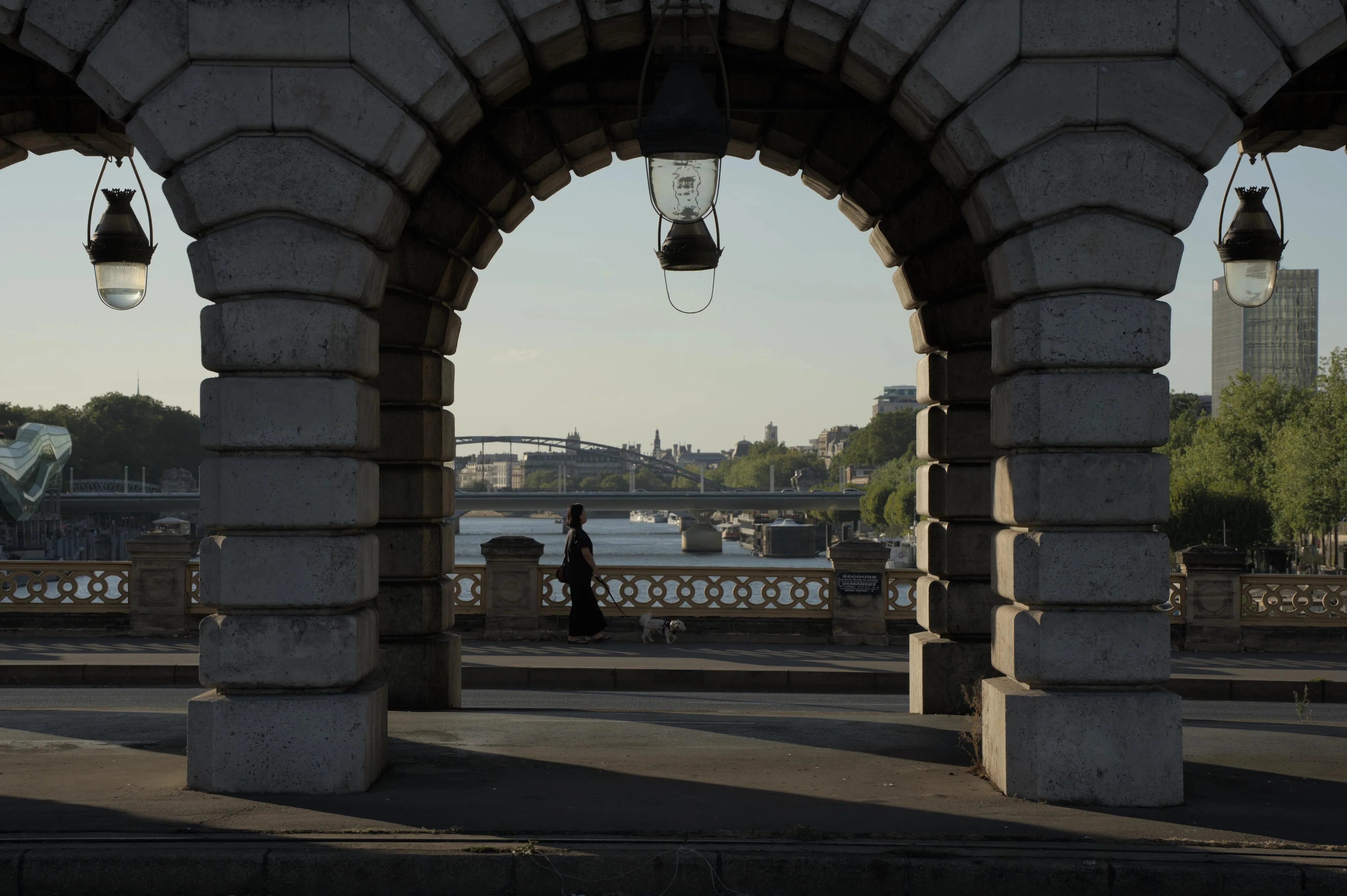 Photograph of a lady walking a small dog on a bridge over a river, seen through a stone arcade built on top of the bridge road. The top of a few historical buildings from central Paris can be seen in the background skyline. The natural light leaves long shadows and has the colors of late afternoon.
