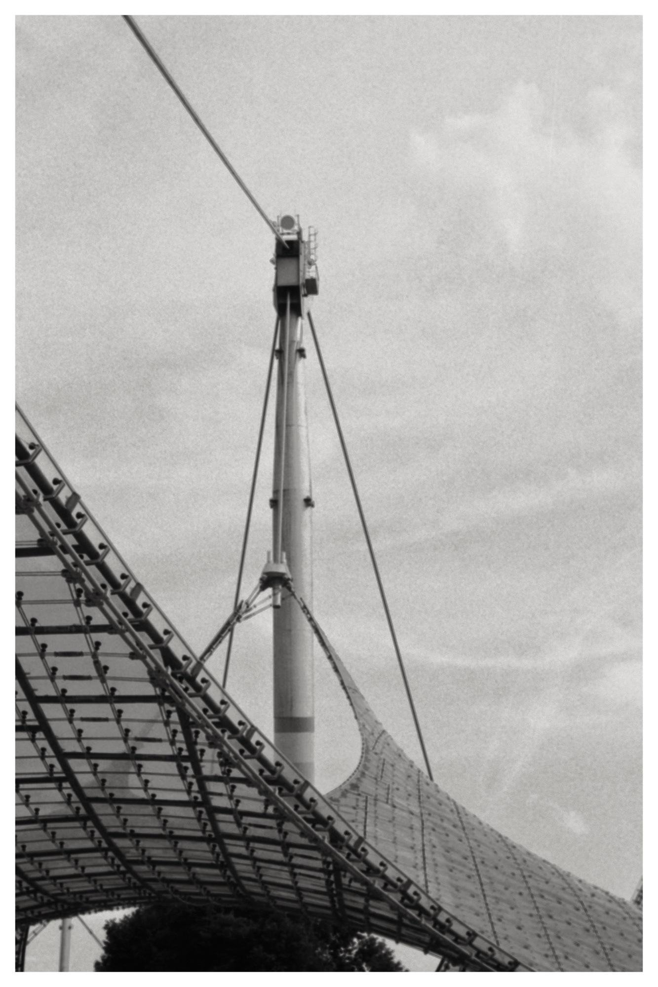 Black-and-white view of a tall mast with cables supporting a curved tensile canopy roof, with the canopy’s metal framework in the lower left and a cloudy sky in the background.