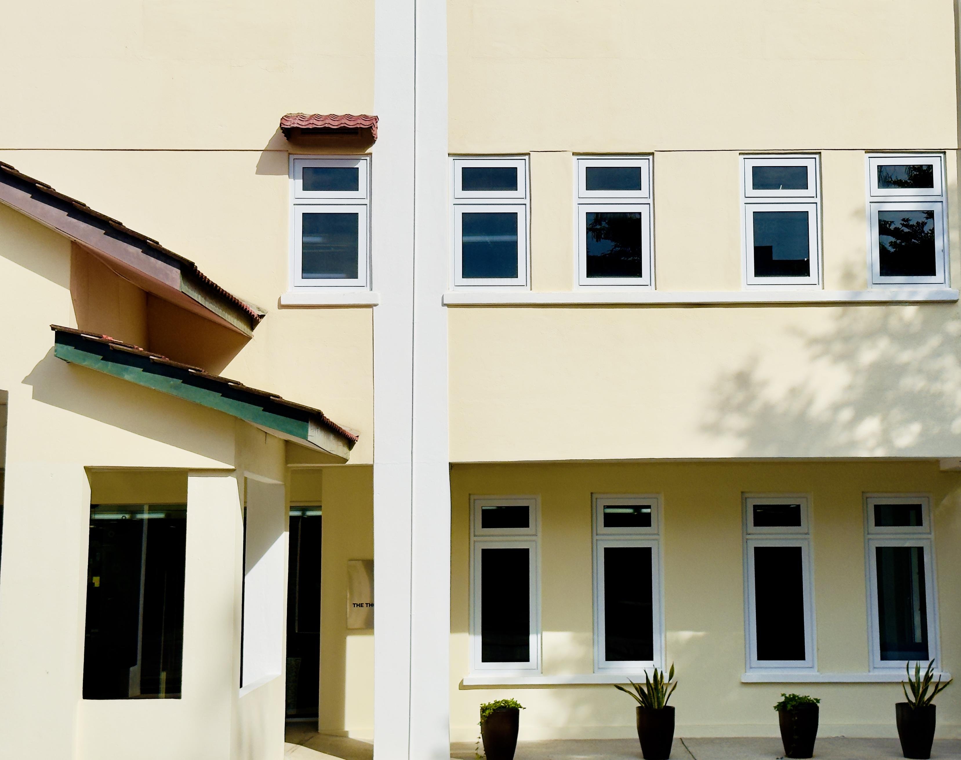 The image shows part of a building with a beige exterior. There are two floors visible, each with several white-framed windows. The top floor has four windows, while the bottom floor has three. There are small potted plants placed in front of the lower windows, and the building features a section of sloped roof with roof tiles. The overall design is modern and simple.