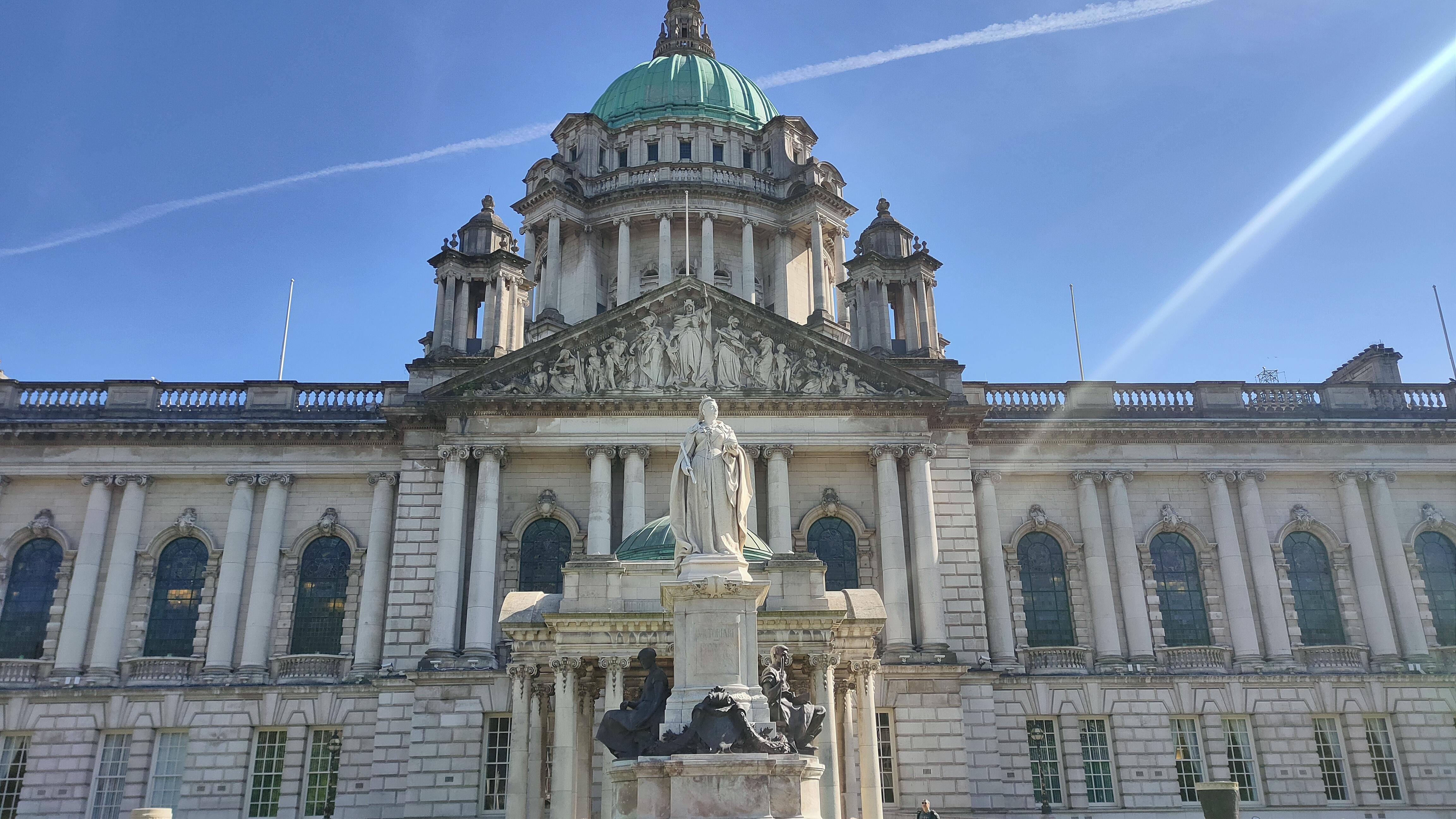 The image shows a grand building with a large dome and classical architectural elements, including columns and statues. There are sculptures and intricate detailing on the facade. 