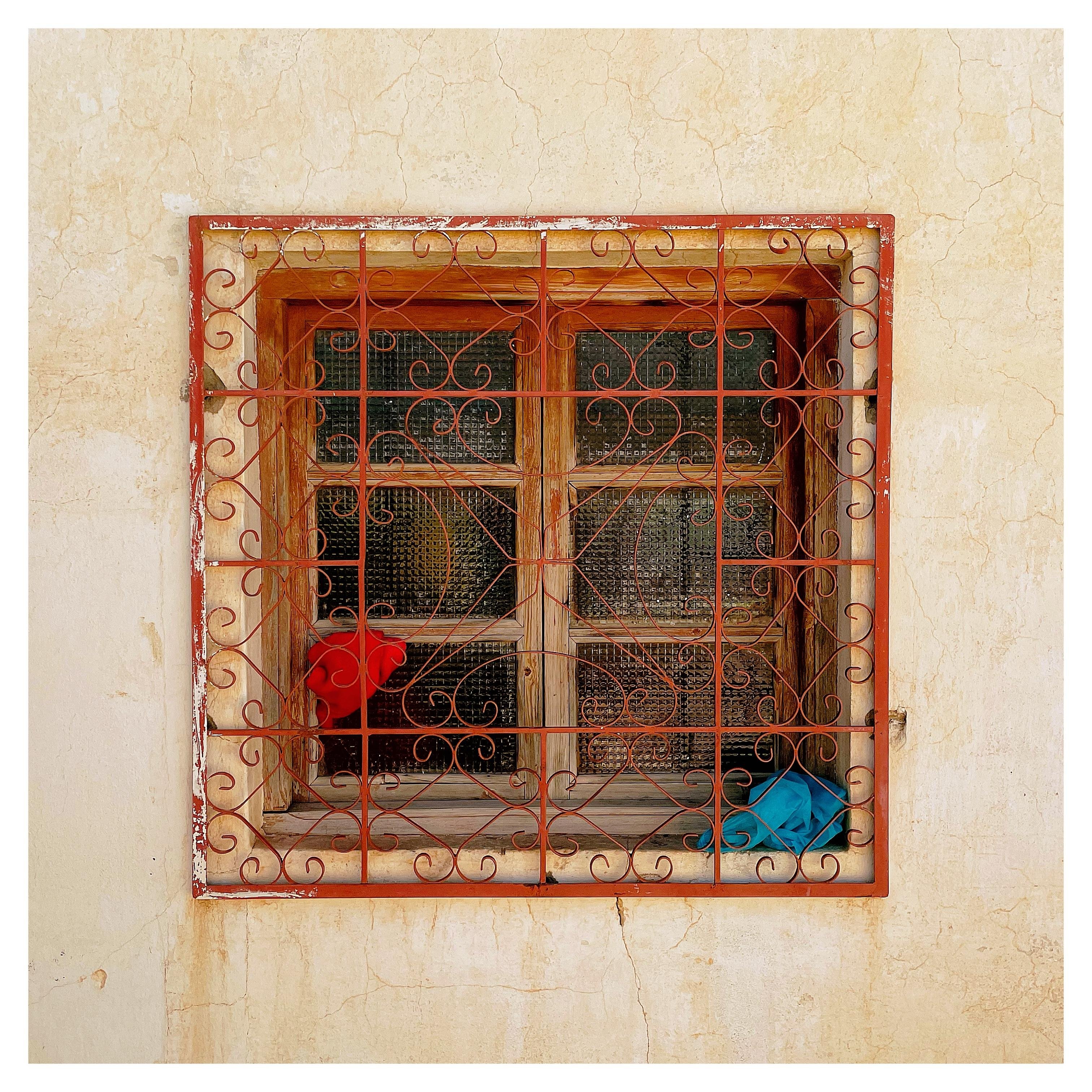 A wooden window with six panes in a wall, covered by a red wrought-iron grille with wavy flourishes; on the left window sill lies a red object, on the right a blue cloth.