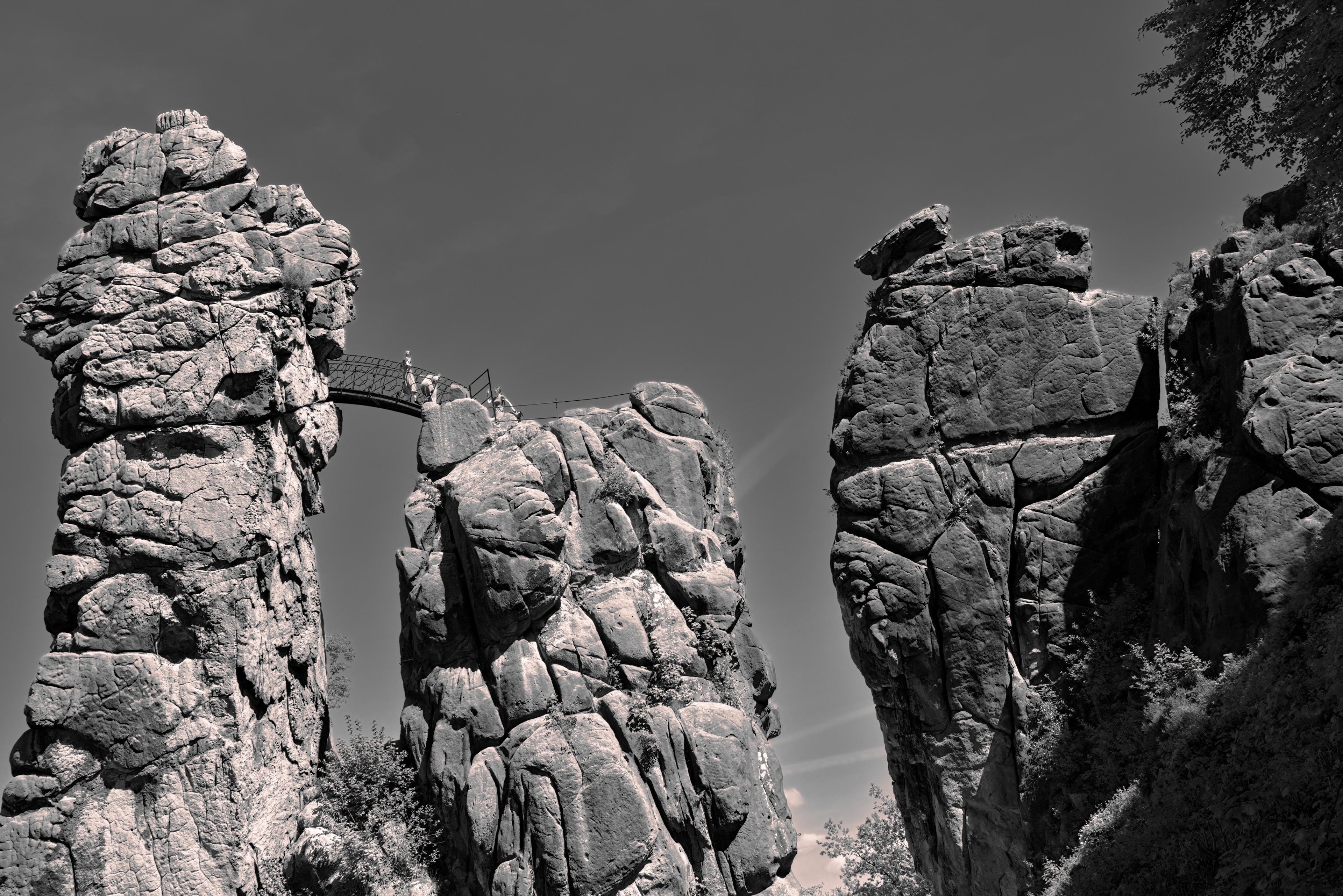The image shows a landscape with large, rugged rock formations connected by a footbridge. These rock formations appear to be natural cliffs or pillars, possibly part of a scenic outdoor location. The sky is mostly clear, suggesting good weather.