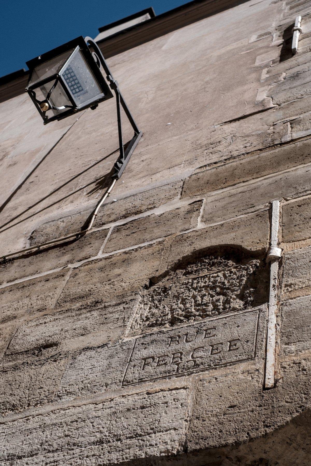 Photo verticale. Contre plongée sur un vieux mur en pierre dans lequel on voit gravé un contour de plaque de nom de rue avec des reliquats d’un nom de rue qui était gravé et « effacé » à coup de dégradations. Juste en dessous, un cadre rectangulaire simple est gravé avec dedans gravé en lettres capitales « RUE PERCEE ». En haut à gauche, un lampion est accroché au mur et on voit un bout de ciel bleu