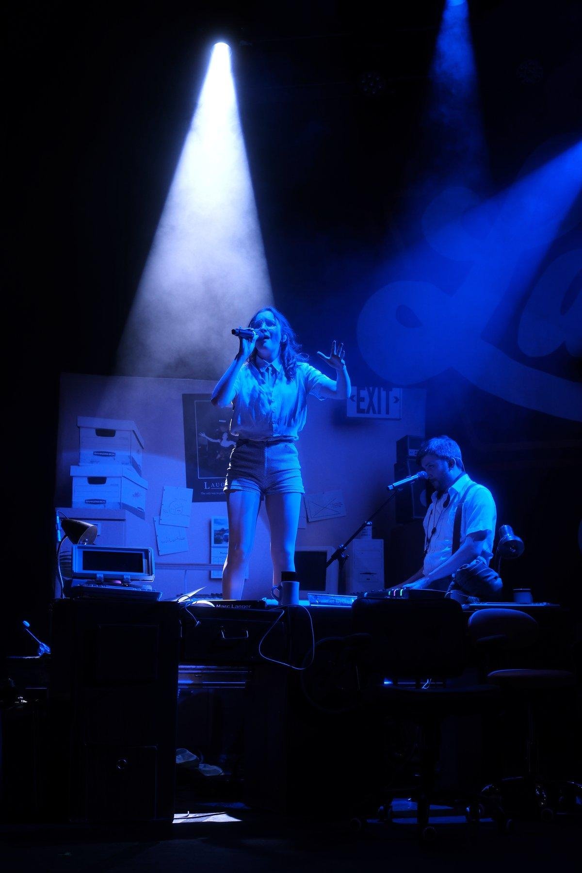 Photo verticale. Gracie Lawrence debout sur un bureau chantant avec une forte prestance. La scène est toute de bleu. Un autre musicien est derrière à droite, jouant du clavier