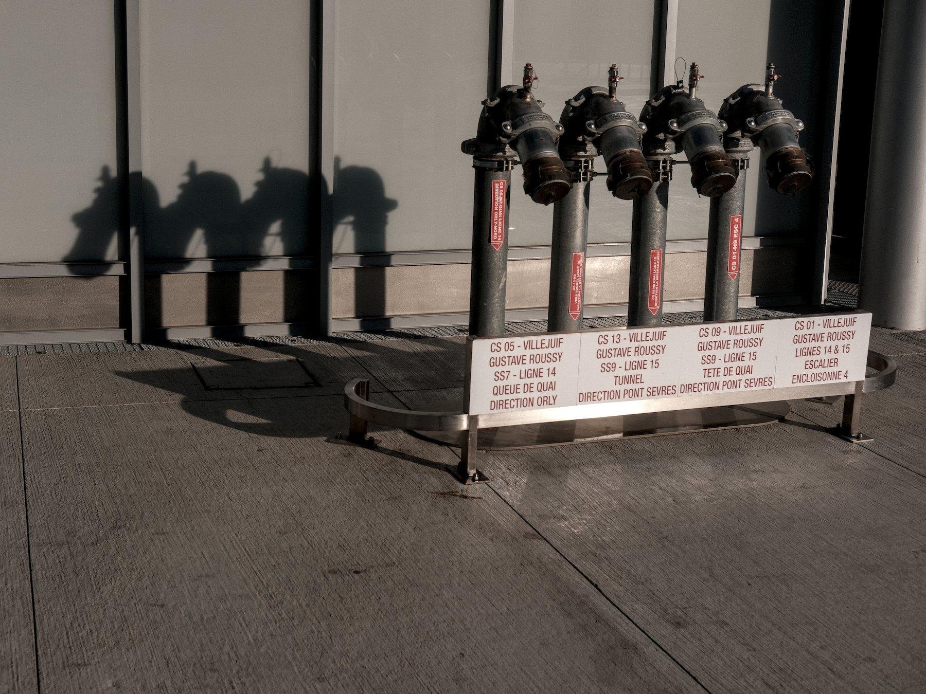 Photo horizontale. 4 tuyaux fermés pour branchement de lance de pompier je suppose, qui descendent dans le sol, avec des étiquettes rouges collées donnant vers quoi ça va et une pancarte blanche à écritures rouge devant. Derrière, leurs ombres est projetée sur le sol et le mur