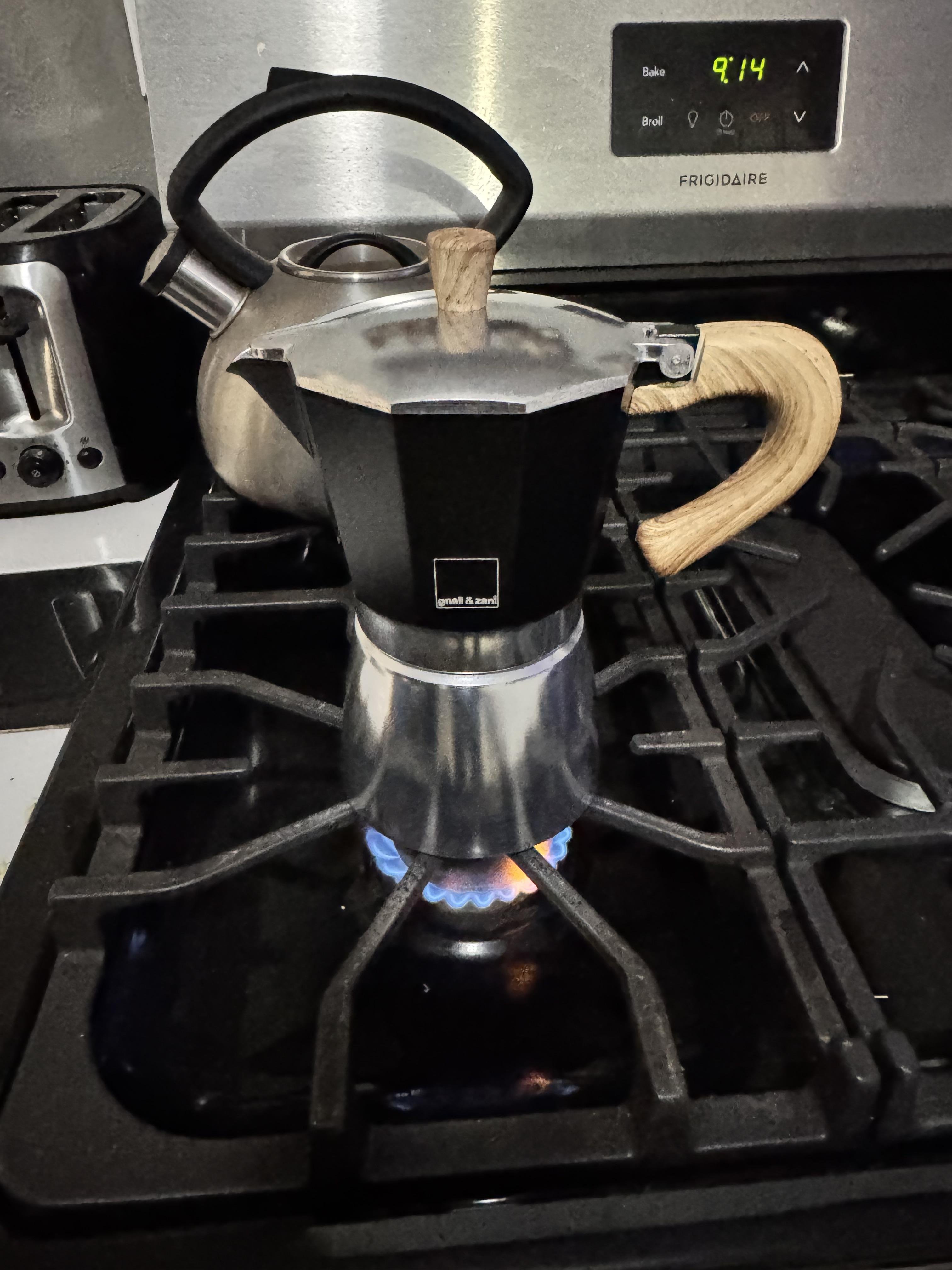 This image shows a stovetop espresso maker, also known as a Moka pot, sitting on a gas stove burner with a visible blue flame underneath it. There’s also a kettle in the background and a toaster on the left. The setting appears to be a kitchen, and the clock on the oven reads 9:14.