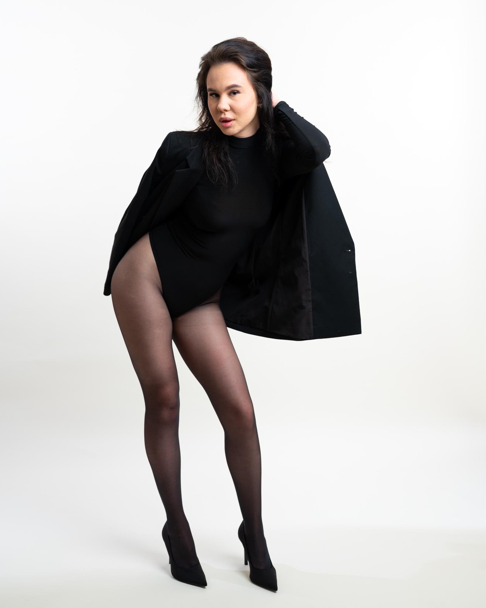 Woman wearing a black leotard, sheer tights, and high heels, with a black jacket draped over her shoulder, standing against a plain white background. Her head is tilted slightly, and one hand holds the jacket while the other rests on her hip.