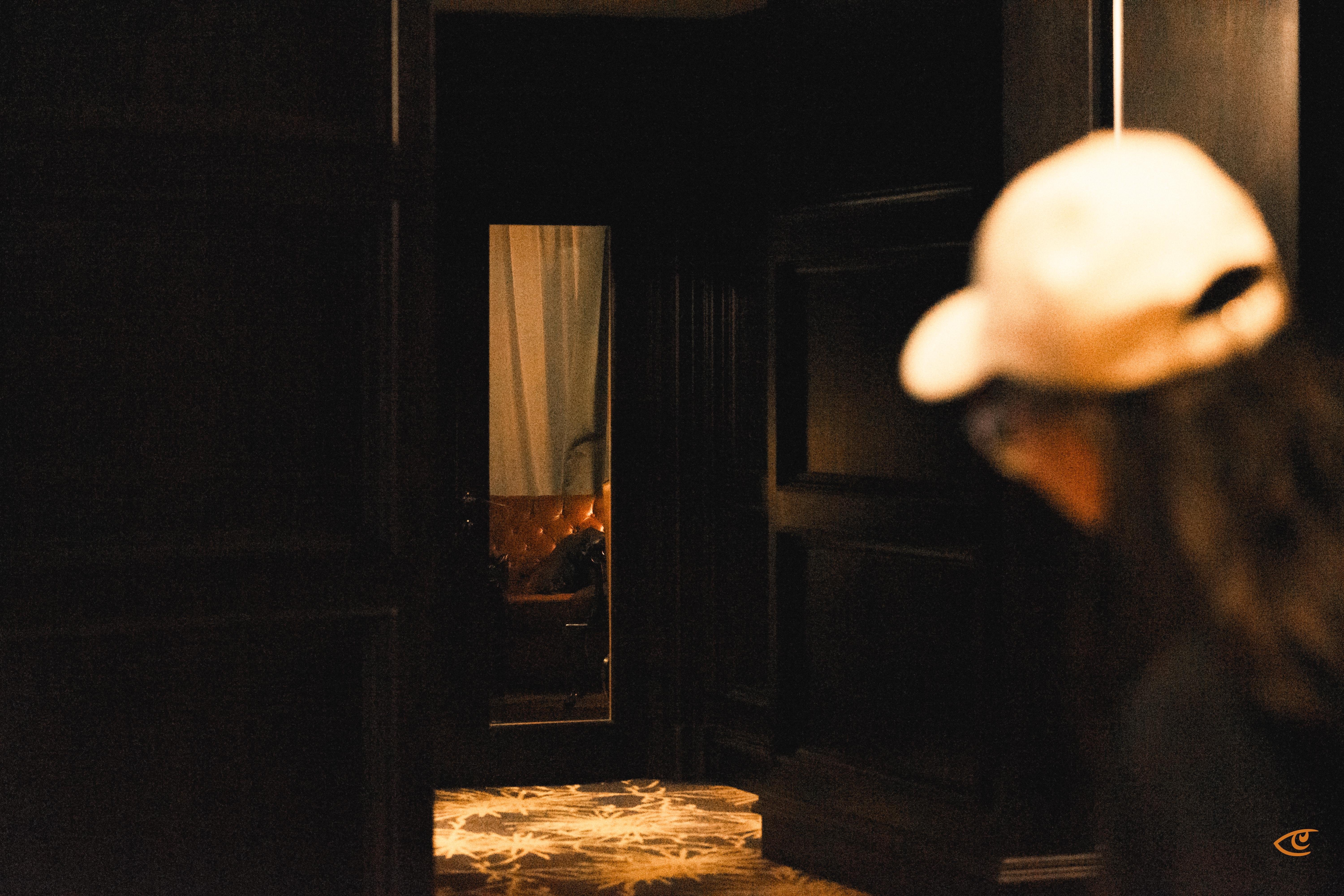 Person in a white baseball cap, seen from behind and out of focus on the right, facing a dim hallway with dark paneled walls and a narrow doorway revealing a lit room with a tufted leather chair; patterned light falls on the floor in the foreground.