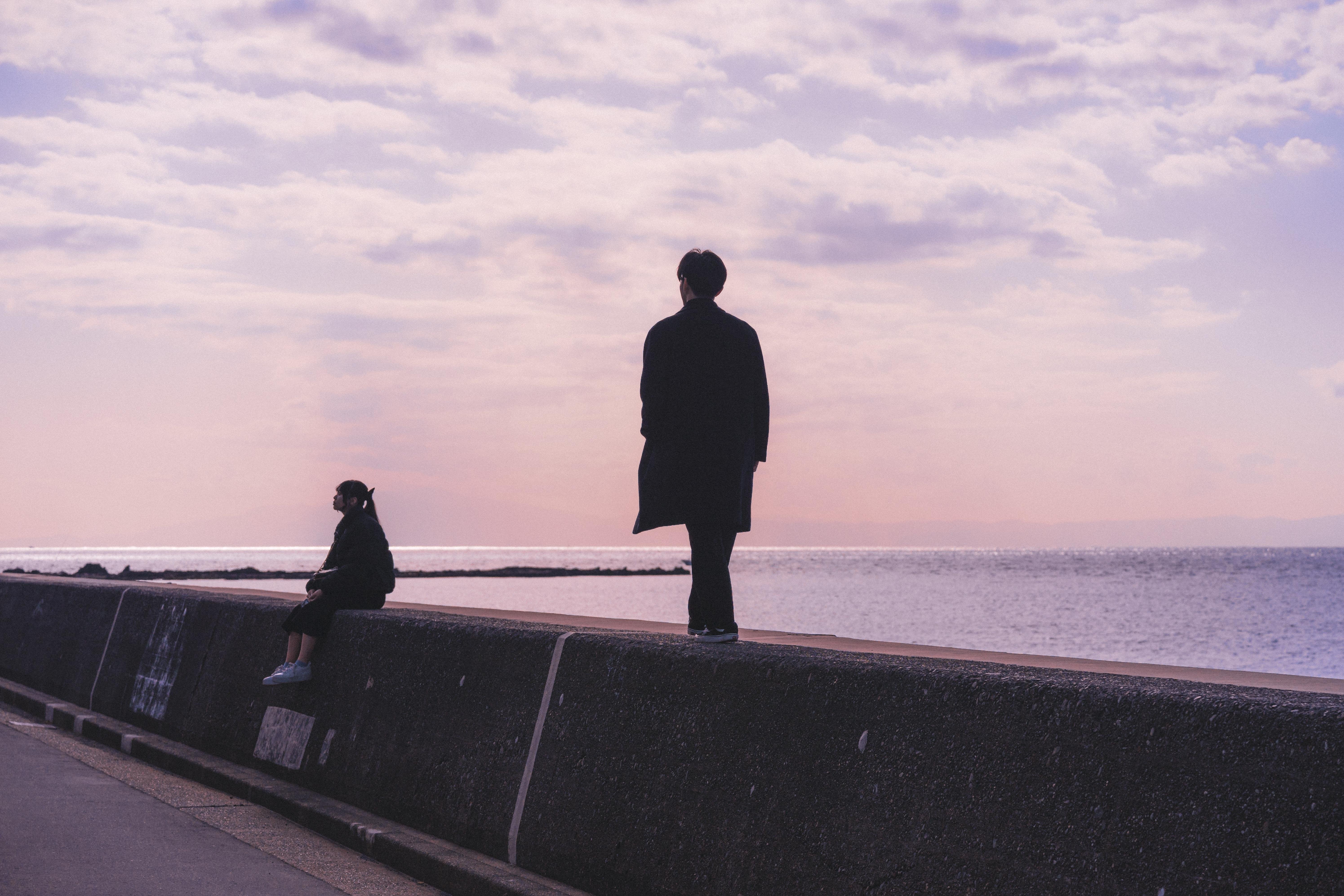 The image depicts two silhouetted figures against a twilight sky over an expansive body of water. The larger figure stands on what appears to be a sea wall or breakwater, dressed in dark clothing with their back turned towards us, gazing out at the horizon where the light pinkish hues meet the darker tones of the evening ocean. Beside them, seated and slightly smaller due to perspective, is another person wearing lighter colors; this figure's posture suggests contemplation as they look away from the water. The environment exudes a quiet atmosphere with no immediate signs of activity or movement around these two figures.