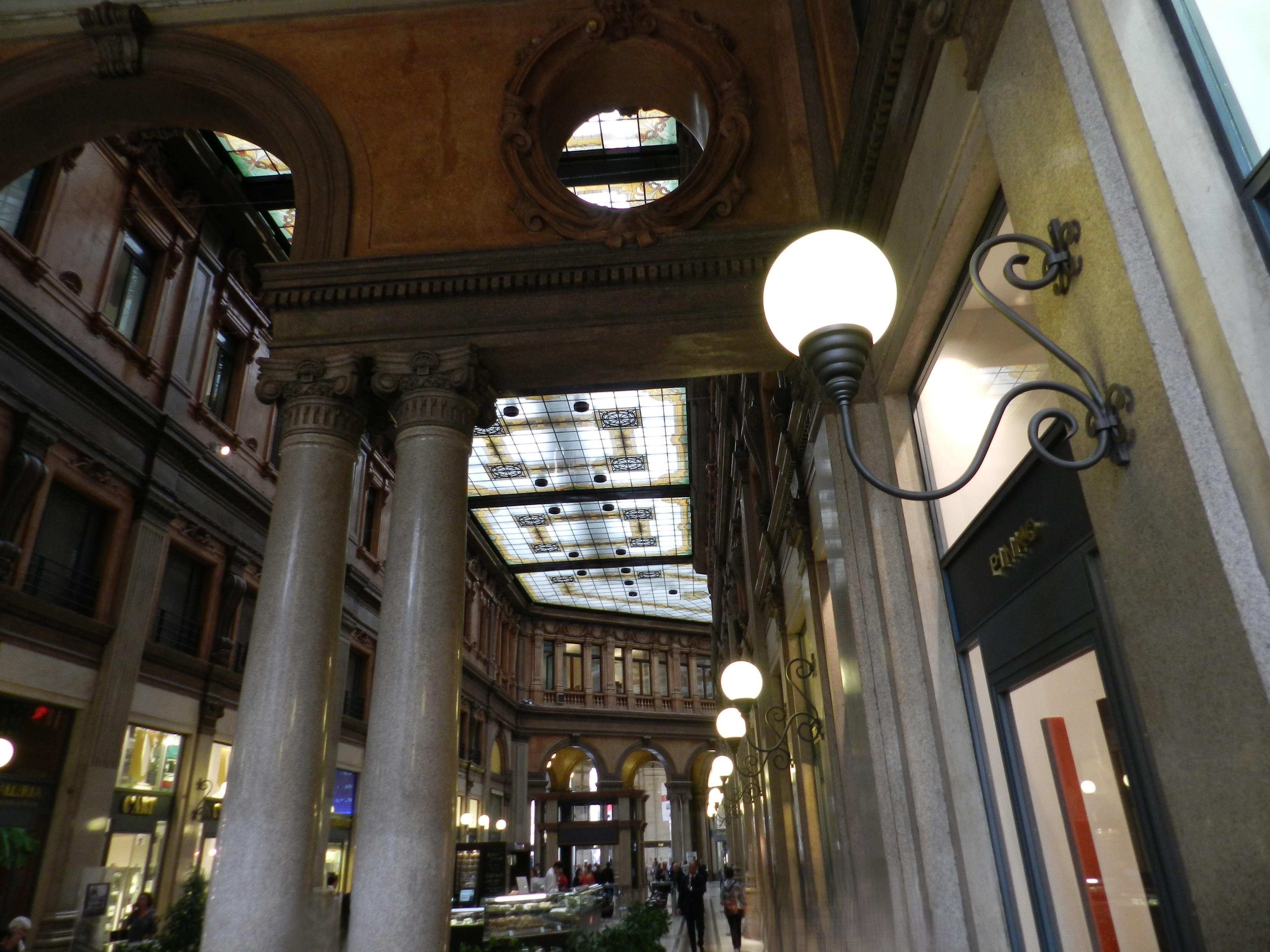 Interior scene of the Galleria Sciarra in Rome, Italy, showing its richly decorated late-19th-century architecture. The image captures a corridor lined with tall, polished stone Corinthian columns supporting an elaborate archway with circular ornamental niches above it. The ceiling is a stunning stained-glass skylight, composed of rectangular panels bordered in warm yellows and greens, with intricate black wrought-iron rosettes that allow soft daylight to filter through. On the right wall, elegant swan-neck metal sconces hold glowing globe lamps, illuminating the walkway and adding a warm contrast to the cool daylight. Shops with refined signage and window displays line the lower level, while the upper floors feature carved window frames, decorative moldings, and a warm terracotta palette. In the distance, another archway opens into the central hall of the gallery. Built originally for the Treves publishing house, the Galleria Sciarra is known for its Art Nouveau details and tranquil, museum-like ambiance.