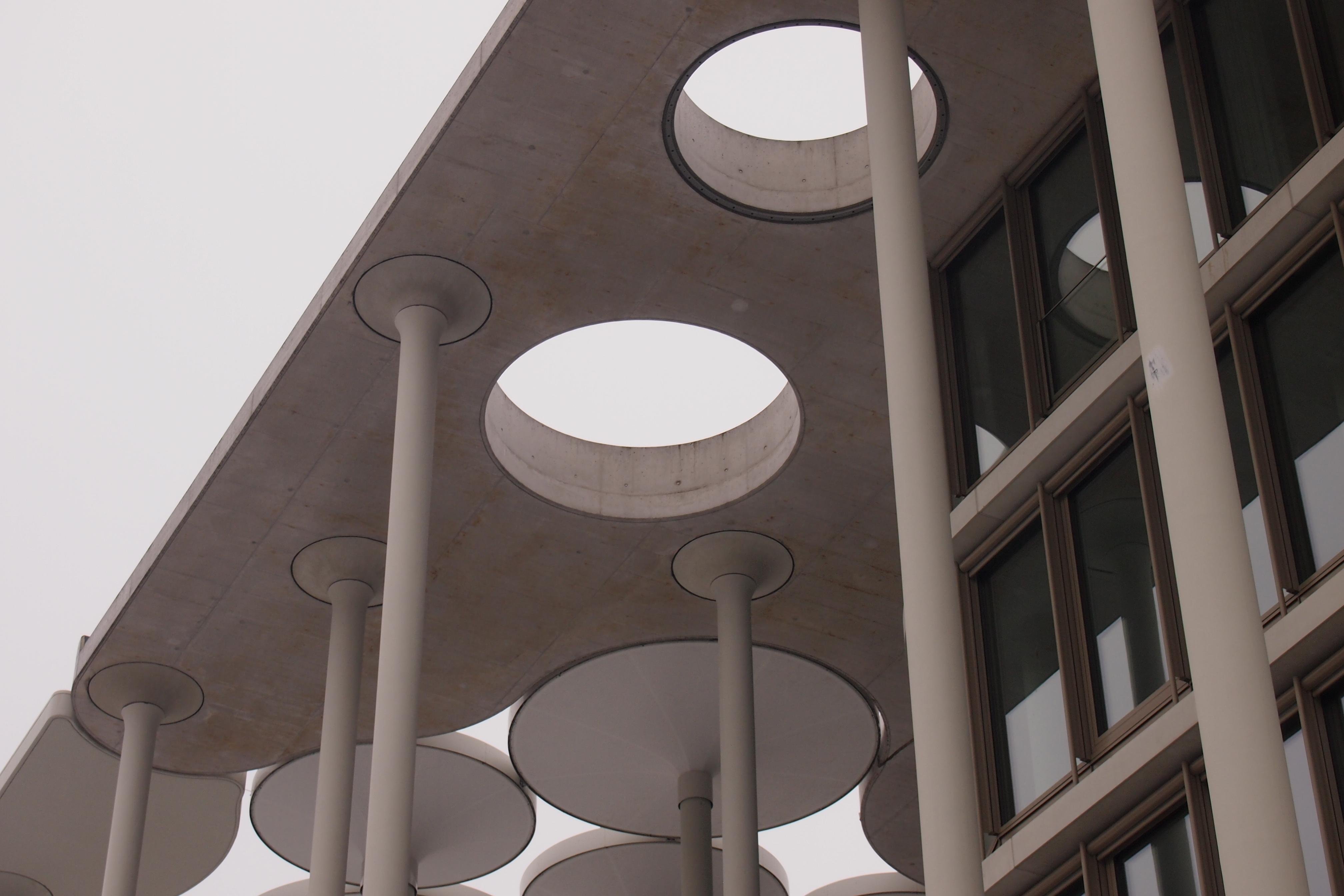 Foto im Querformat.
Blick von Schräg unten auf die oberen Stockwerke und das Dach eines Bürogebäudes.
Rechts befinden sich Fenster aus drei Etagen, links einige Betonsäulen, die nach oben in einzelne Kreise bzw. eine geschlossene Betondecke mit runden Öffnungen übergehen.
Der Himmel in der linken oberen Ecke ist in einem sehr hellen Grau.