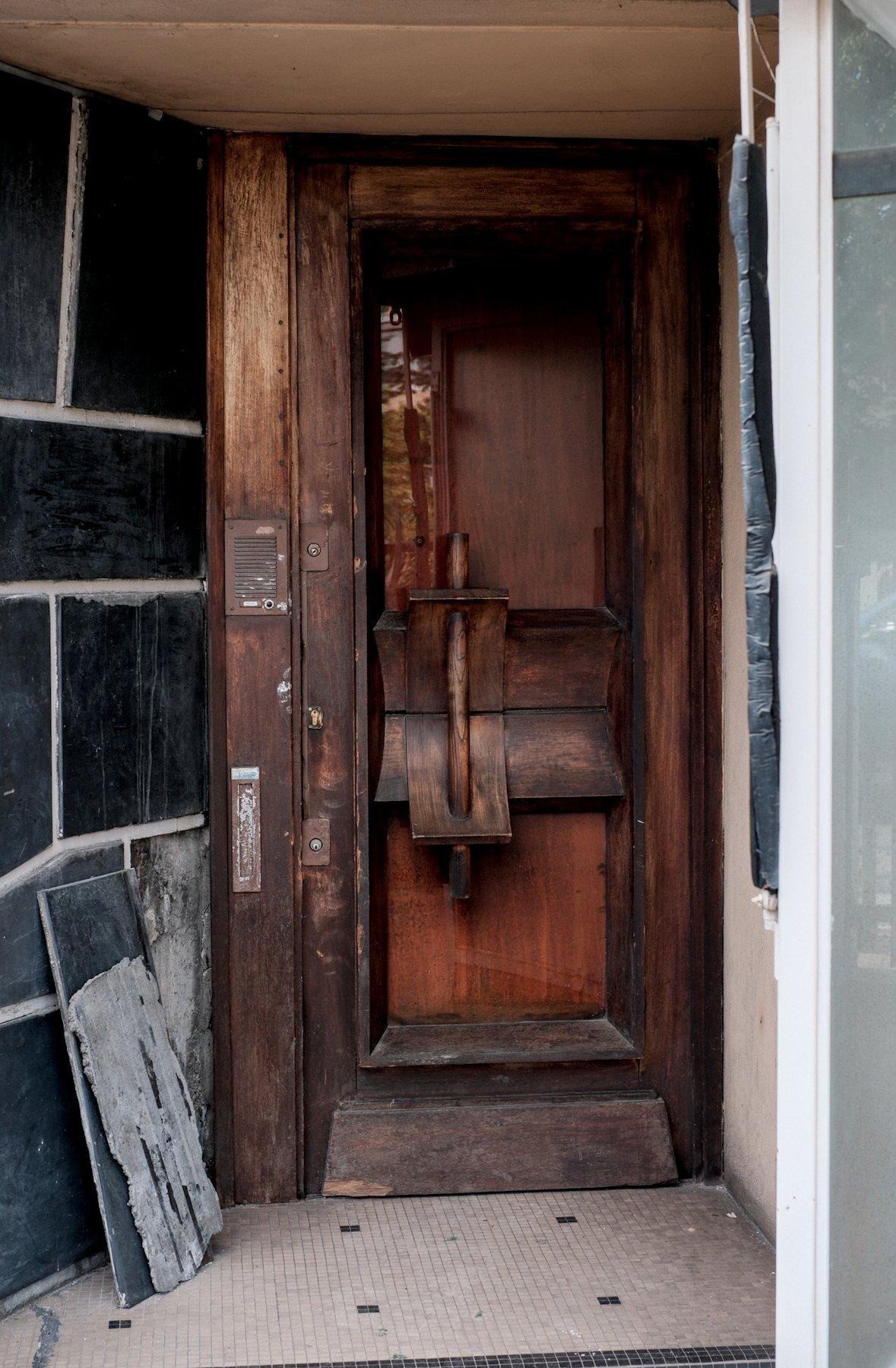 Photo verticale. Une vieille porte en bois avec une poignée sortant de l’ordinaire, du même bois. Le palier a un mur avec de grande plaques de céramiques bleu sombre, dont une est détachée et posée au sol, contre le mur