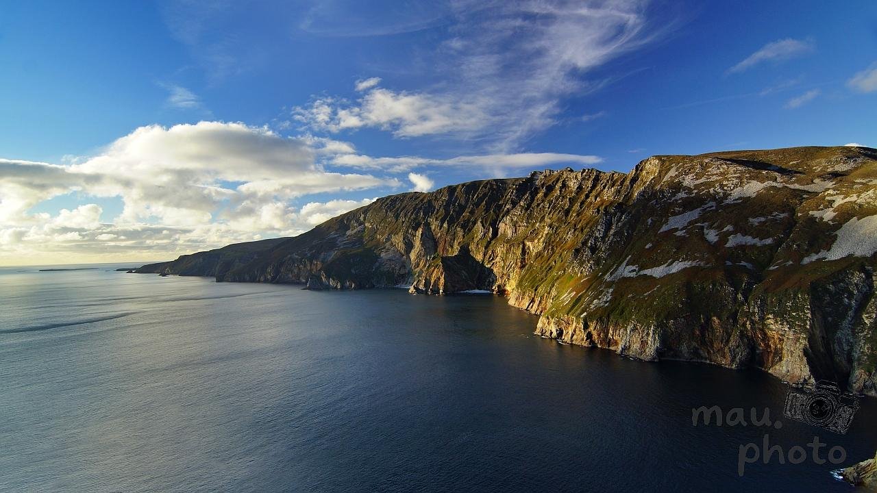 Wide view of rugged sea cliffs curving along a coastline, with dark blue water below and a partly cloudy blue sky above; patches of grass and light snow cover the cliff tops.