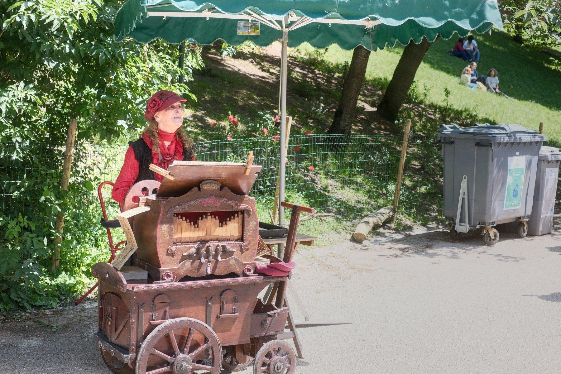 Photo horizontale. Portrait d’une dame habillée en rouge, jolie casquette rouge avec une fleur sculpté dans le tissu sur le côté, Un gilet noir pour le contraste. Elle joue d’un orgue de barbarie en bois et à roulettes. On voit sur la gauche, le papier qui sort de la machine et on voit un bout du pendant à droite. Le tout se trouve sous un large parasol rectangulaire vert